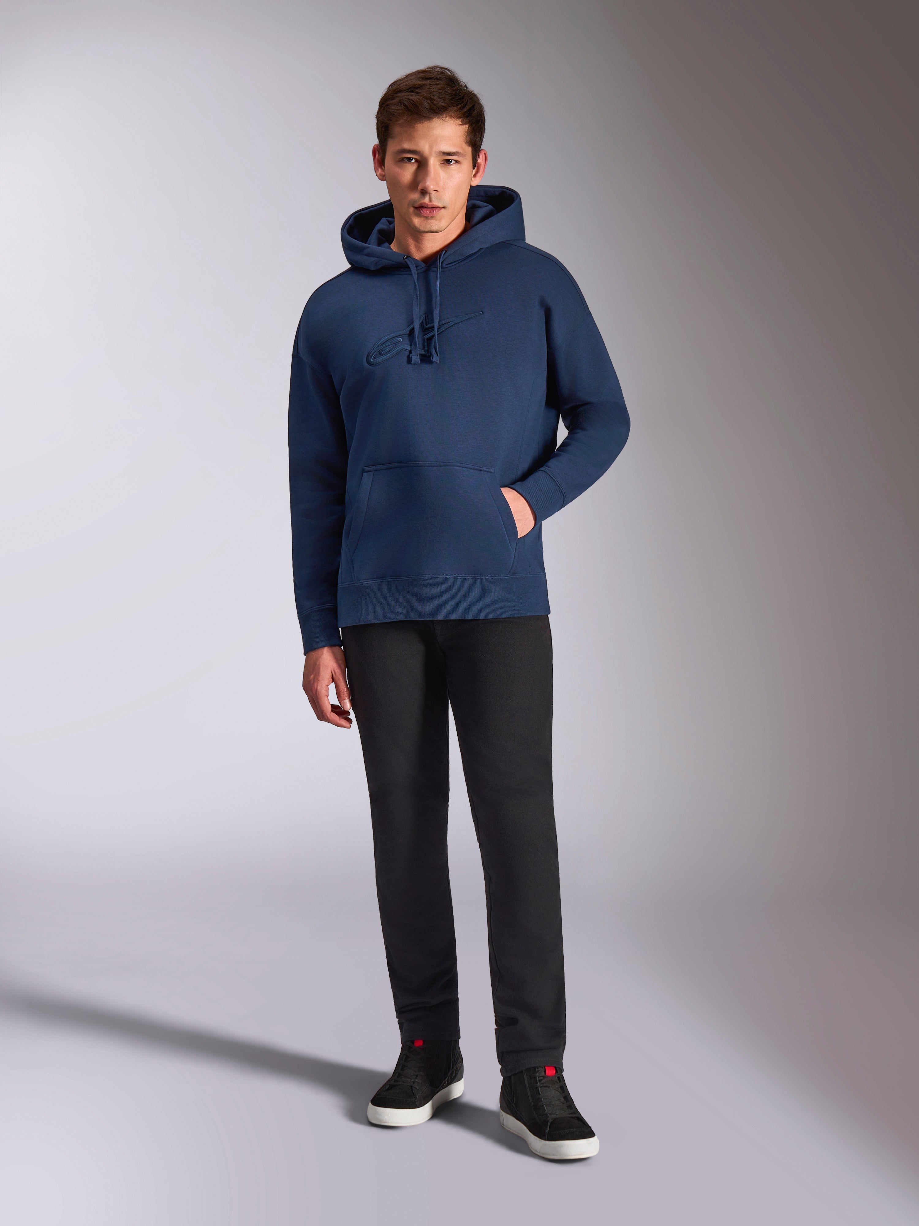 Alpinestars Emergent Oversized Hoodie, marineblauer Fleece-Kapuzenpullover, farblich abgestimmte 3D-Logostickerei auf der Vorderseite, getragen von einem Modell mit schwarzer Hose und Sneakern, lässiger Streetwear-Stil.
