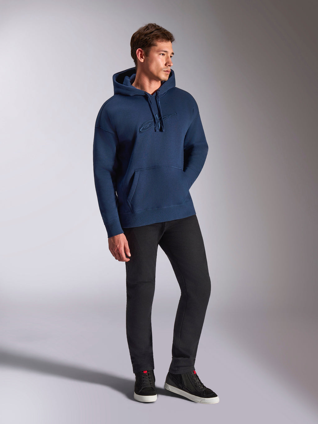 Alpinestars Emergent Oversized Hoodie, Marineblau, getragen von einem männlichen Model mit schwarzer Hose und Sneakern, mit 3D-gesticktem Astars-Logo, Kängurutasche und Kapuze mit Kordelzug, lässige, vom Motorsport inspirierte Streetwear.