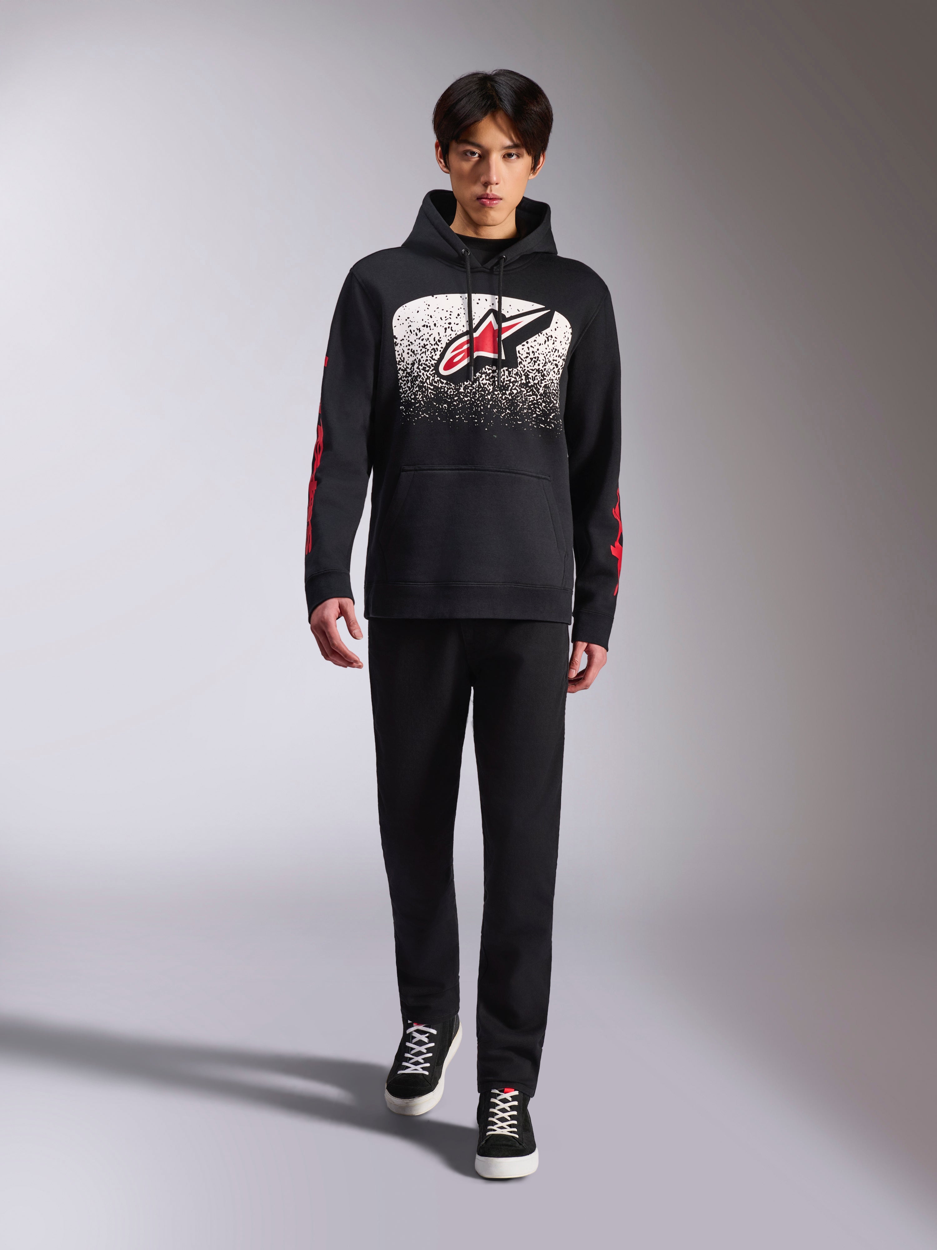 Alpinestars Obsessed Hoodie, schwarzer Kapuzenpullover, großes weißes und rotes Astars-Logo mit MX-Farbverlauf, rote Wortmarken an den Ärmeln, getragen von einem Model in einem komplett schwarzen Freizeitoutfit, motorsport-inspirierte Lifestyle-Bekleidung