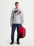 Alpinestars Obsessed Hoodie, Sportswear-Kapuzenpullover für Herren, Zementgrau, Rot und Schwarz, MX-inspirierte Verlaufsgrafiken und Ärmellogos. Gezeigt an einem männlichen Modell, das einen roten Alpinestars Rucksack hält.