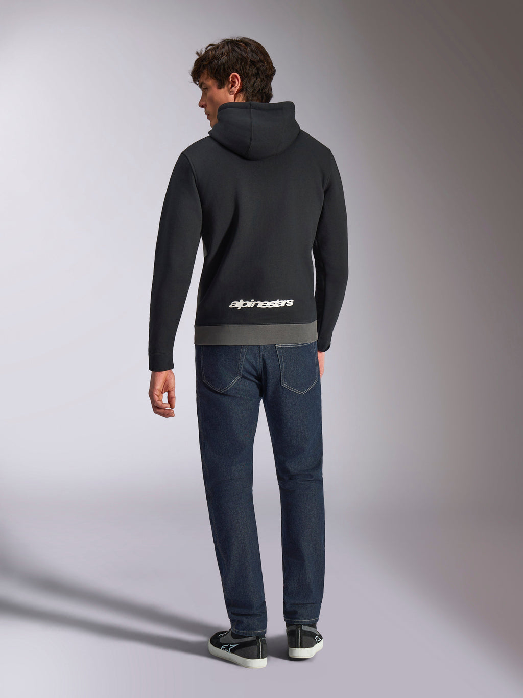 Alpinestars Parallel Hoodie, Rückansicht eines Mannes in einem Fleece-Pullover mit Color-Blocking in Schwarz und Gunmetal, weißes Alpinestars-Wortlogo am unteren Rücken, kombiniert mit dunkelblauen Jeans, lässige Motorsport-inspirierte Streetwear