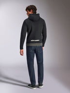 Alpinestars Parallel Hoodie, Rückansicht eines Mannes in einem Fleece-Pullover mit Color-Blocking in Schwarz und Gunmetal, weißes Alpinestars-Wortlogo am unteren Rücken, kombiniert mit dunkelblauen Jeans, lässige Motorsport-inspirierte Streetwear