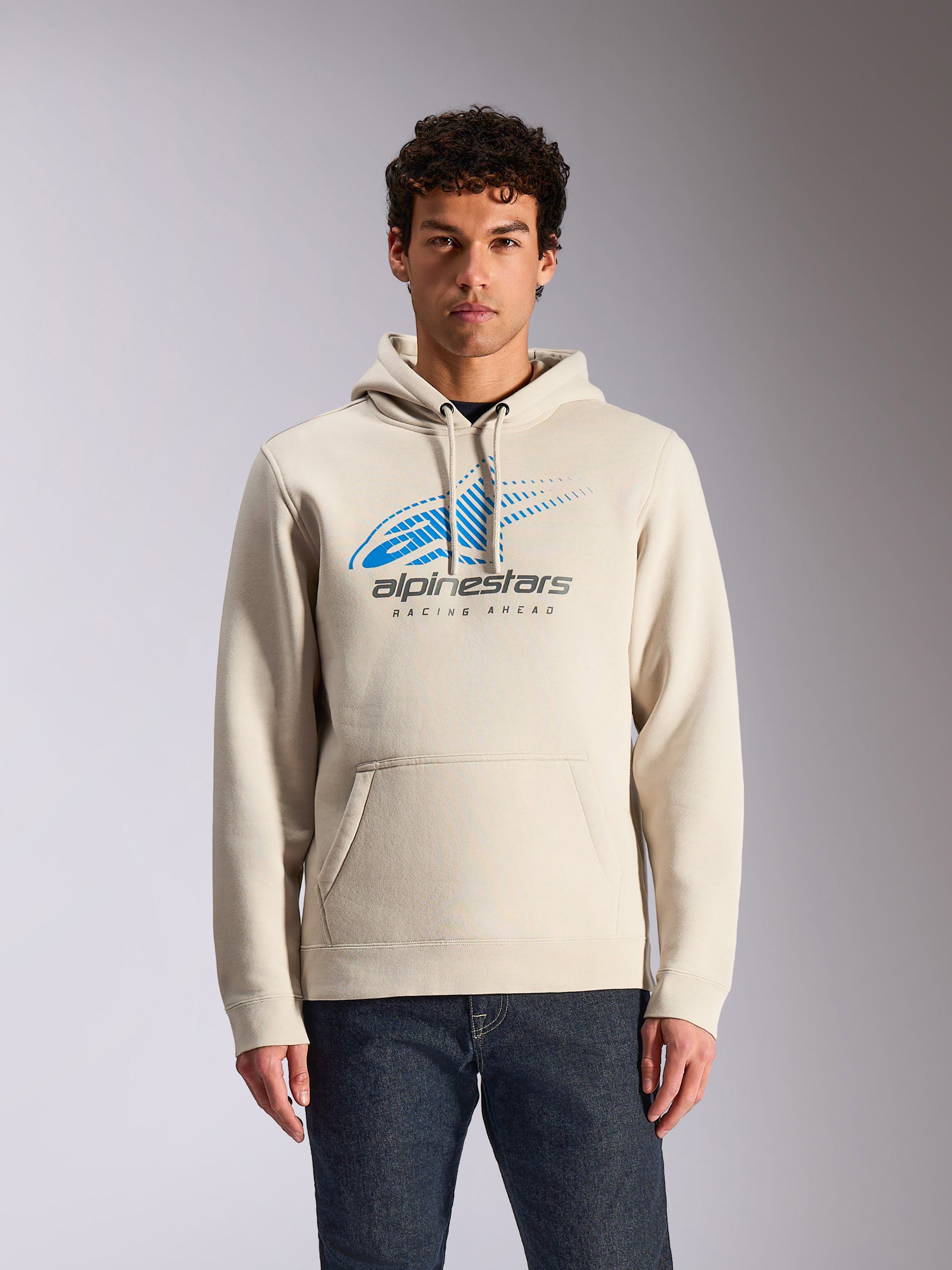 Alpinestars Pulse Hoodie, lässiger Kapuzenpullover in Off-White, blaues und graues Astars Combo-Logo auf der Vorderseite mit Speed-Line-Fade-Grafik, vordere Kängurutasche, von einem Model mit dunkler Jeans getragen, Motorsport-inspirierte Streetwear