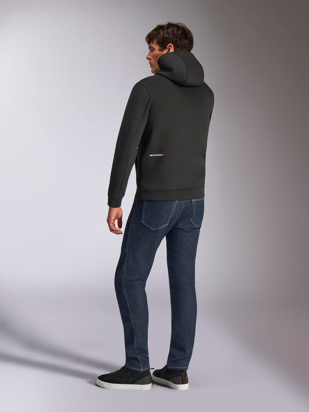 Alpinestars Cypher Hoodie, Black casual zip hoodie, rear view showing high neck hood and white Alpinestars logo on the lower back, worn with dark blue jeans and black sneakers, modern motorsport-inspired design