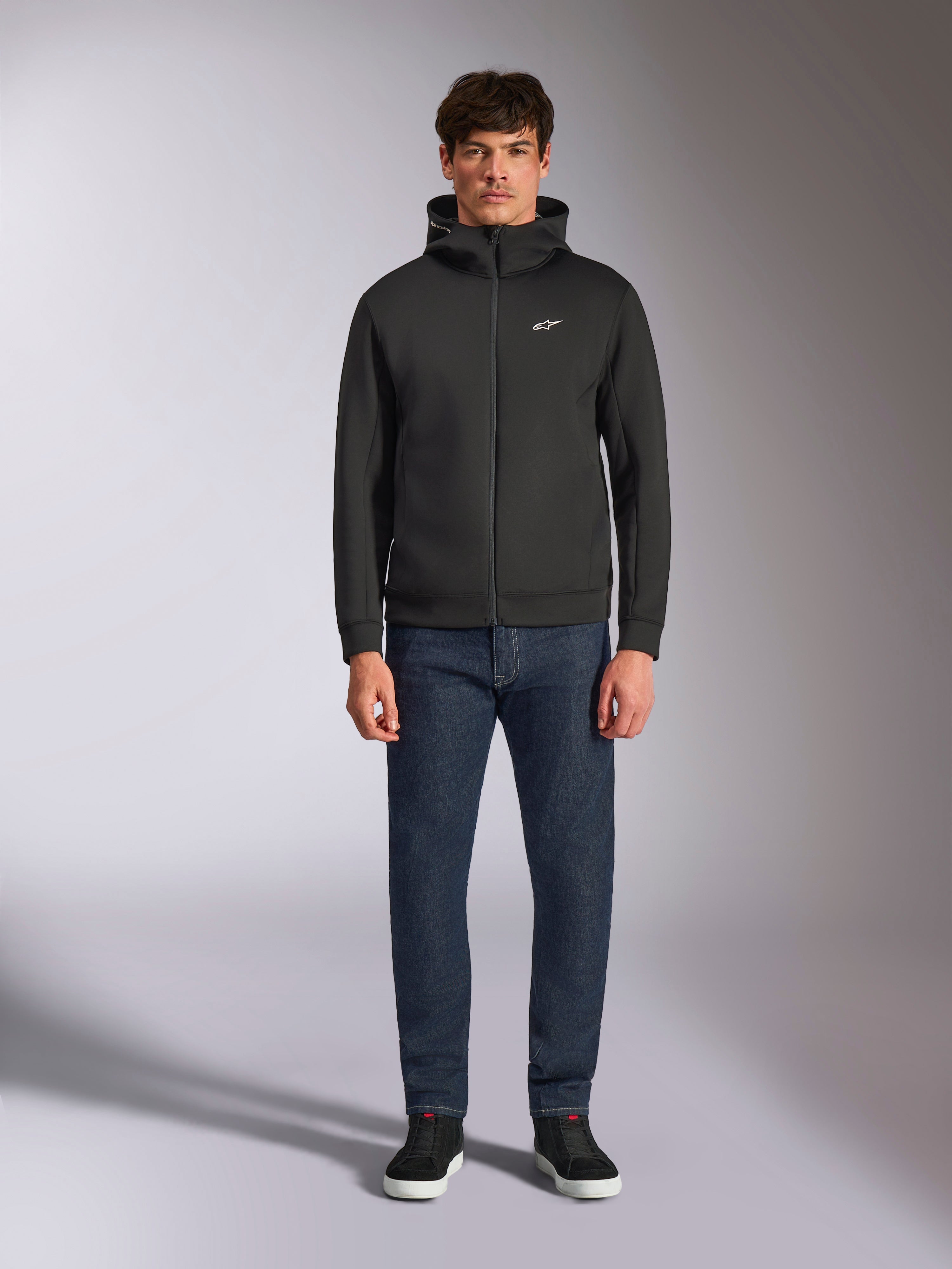 Model wearing the Alpinestars Cypher Hoodie, Black zip-front fleece hoodie, high-neck hood with a small white Astars logo on the chest, paired with dark denim jeans and black sneakers, casual motorsport-inspired streetwear