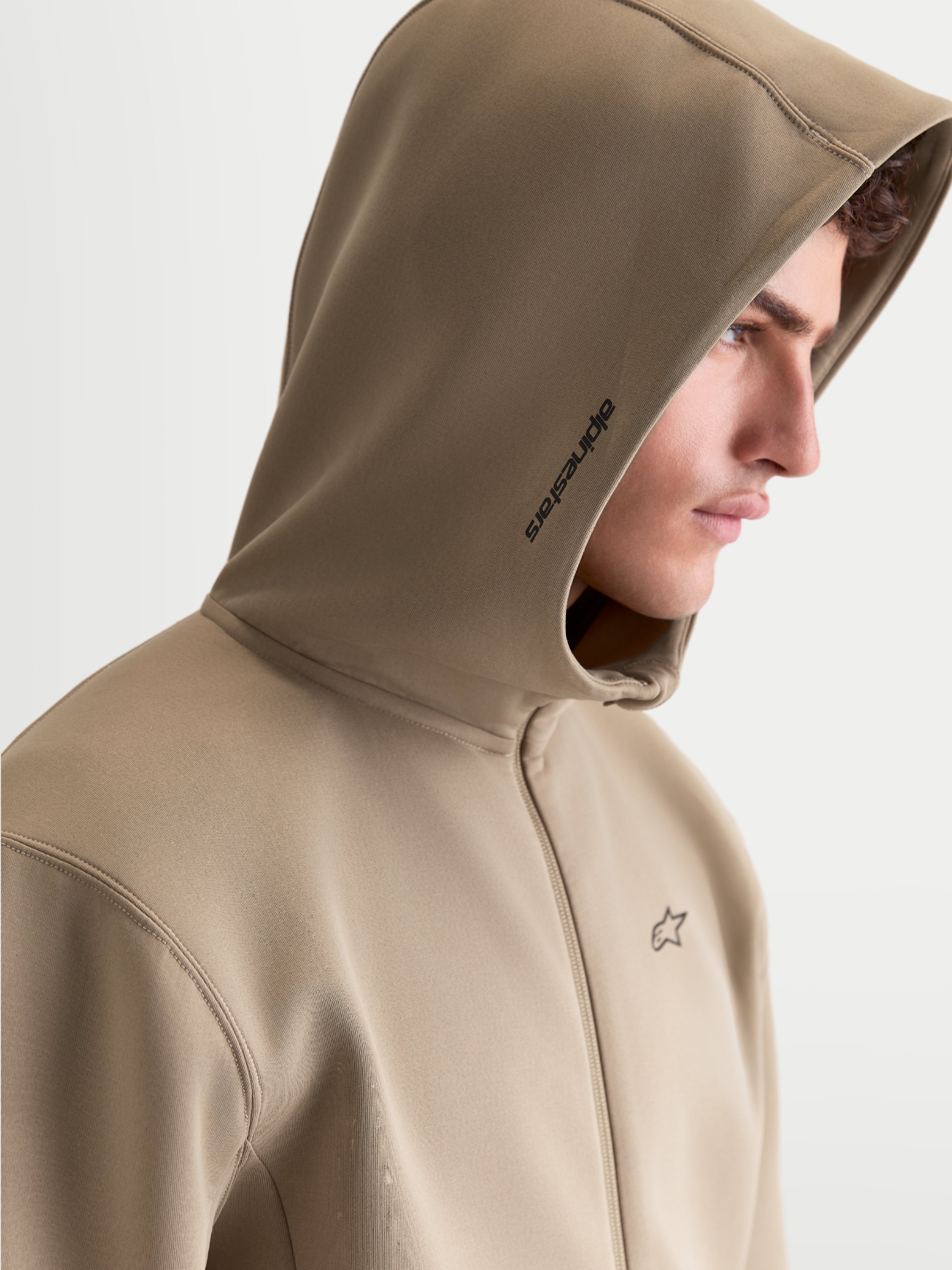 Alpinestars Cypher Hoodie, Sportswear-Kapuzenjacke mit Reißverschluss für Herren, Putty Beige. Profilansicht am Modell mit hochgeschlossener Kapuze inklusive Mesh-Futter, Alpinestars-Branding an der Seite und Logo auf der Brust. Hochwertiges Tri-Blend-Fleece für einen modernen Motorsport-Look.
