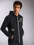Alpinestars Transverse Kapuzenpullover, lässige Sportbekleidung, Schwarz mit weißem Reißverschluss, Kapuze mit Kordelzug, Alpinestars Stern-Logo auf der Brust, „One Goal One Vision“-Grafik, getragen von einem männlichen Model mit dunklen lockigen Haaren