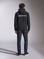 Alpinestars Transverse Hoodie, lässige Motorsport-Streetwear, Schwarz mit weißem Alpinestars-Wortlogo auf dem Rücken, lange Ärmel, Rippbündchen an Saum und Ärmeln, Rückansicht getragen mit dunkler Denim-Jeans