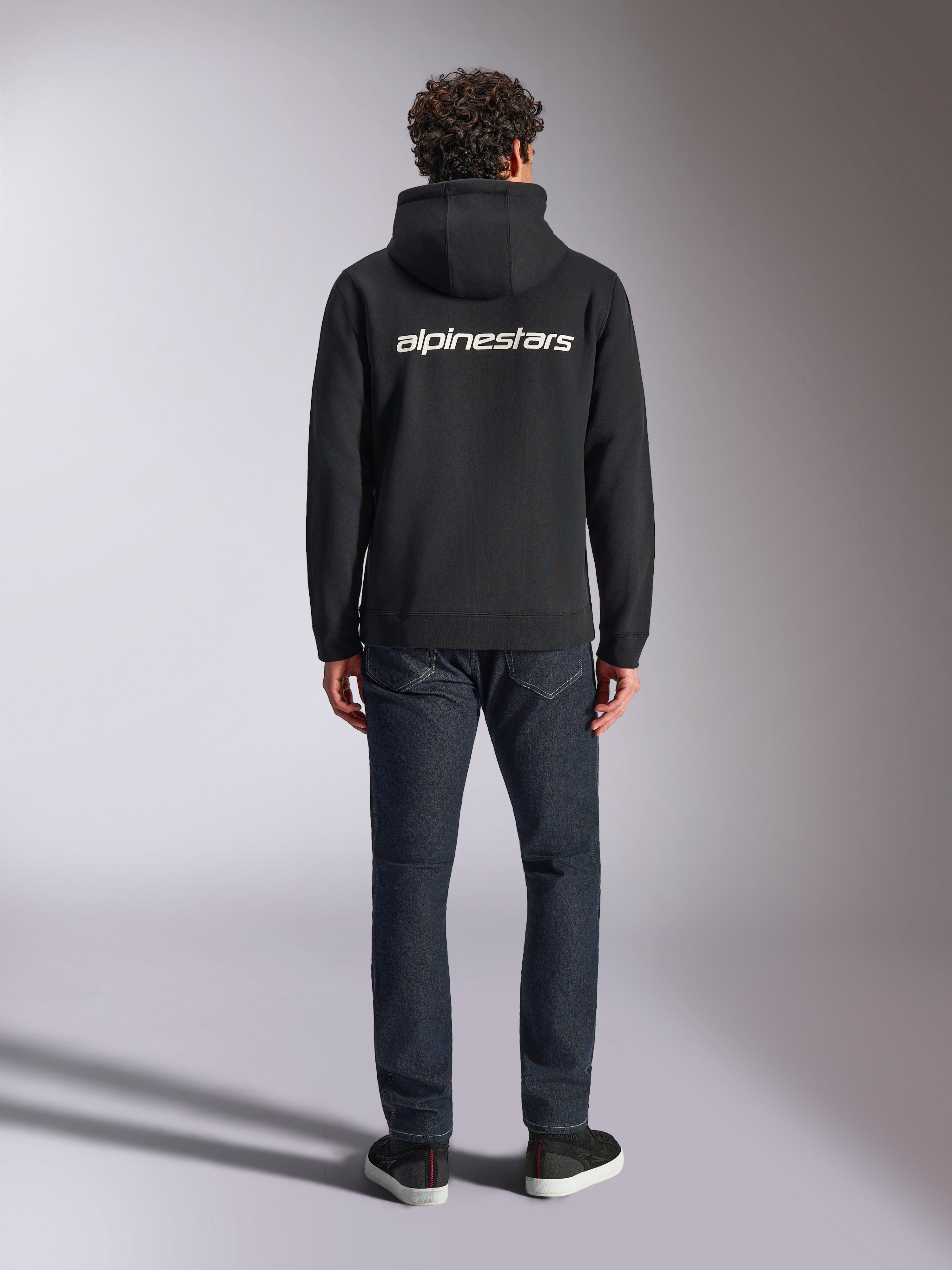Alpinestars Transverse Hoodie, lässige Motorsport-Streetwear, Schwarz mit weißem Alpinestars-Wortlogo auf dem Rücken, lange Ärmel, Rippbündchen an Saum und Ärmeln, Rückansicht getragen mit dunkler Denim-Jeans