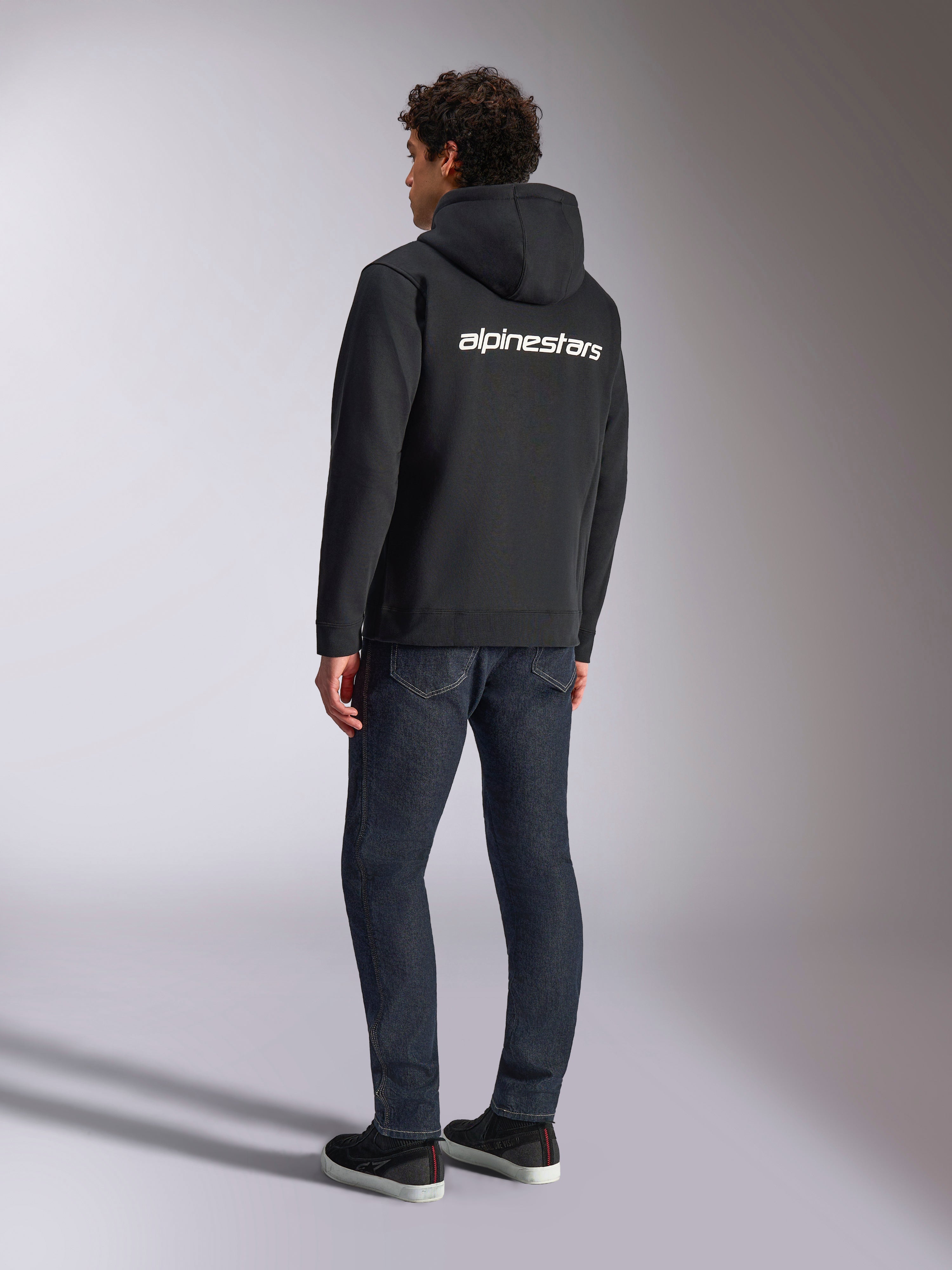 Alpinestars Transverse Hoodie, schwarzer Kapuzenfleece mit Reißverschluss, Rückansicht mit weißem Alpinestars-Schriftzug über den Schultern, kombiniert mit dunkelblauen Jeans und schwarzen Sneakern, lässige, vom Motorsport inspirierte Streetwear