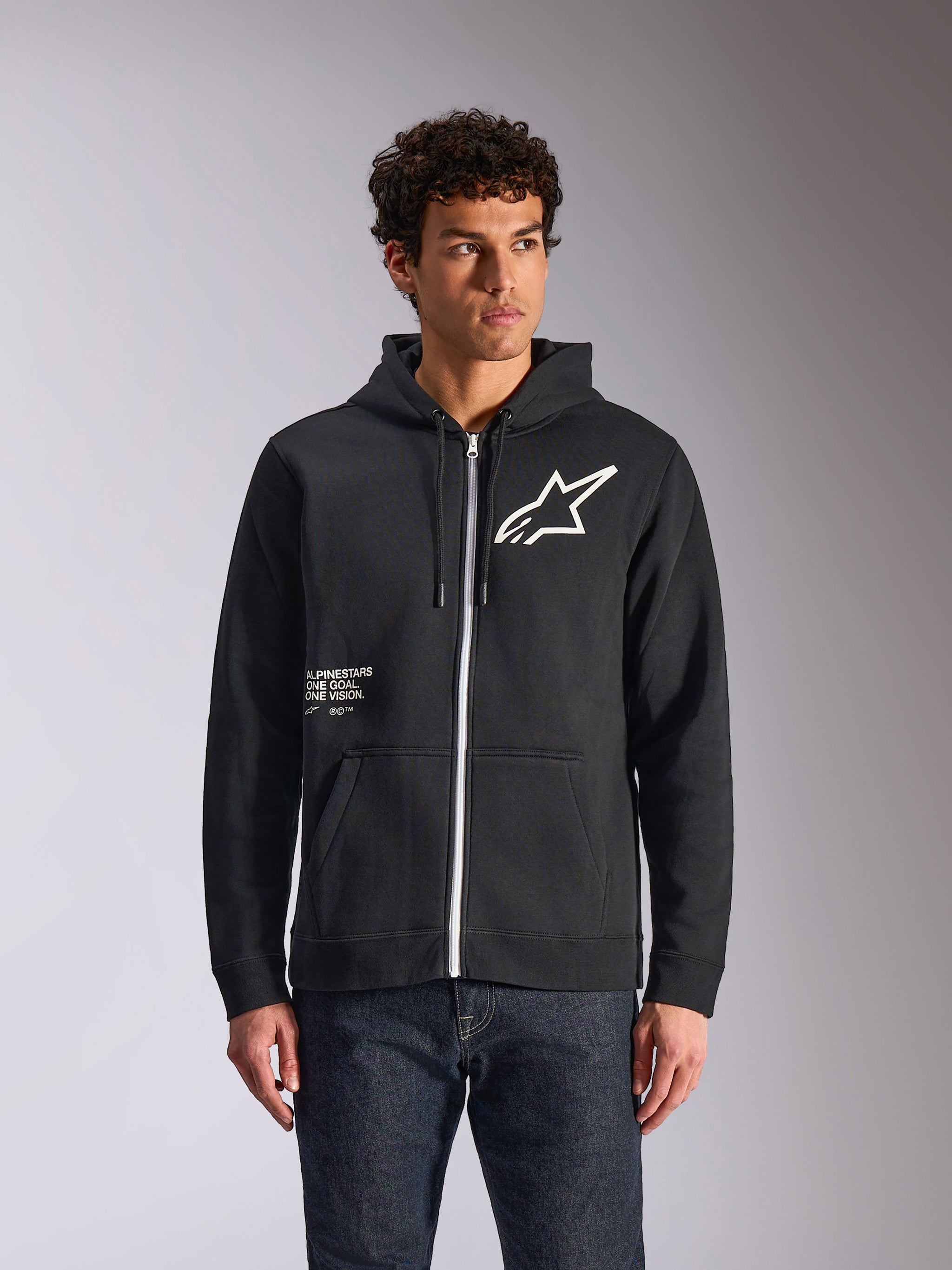 Alpinestars Transverse Hoodie, Black zip-front hooded fleece, featuring white Astars logo and One Goal graphic on the front, worn by a model with dark jeans, casual motorsport-inspired streetwear