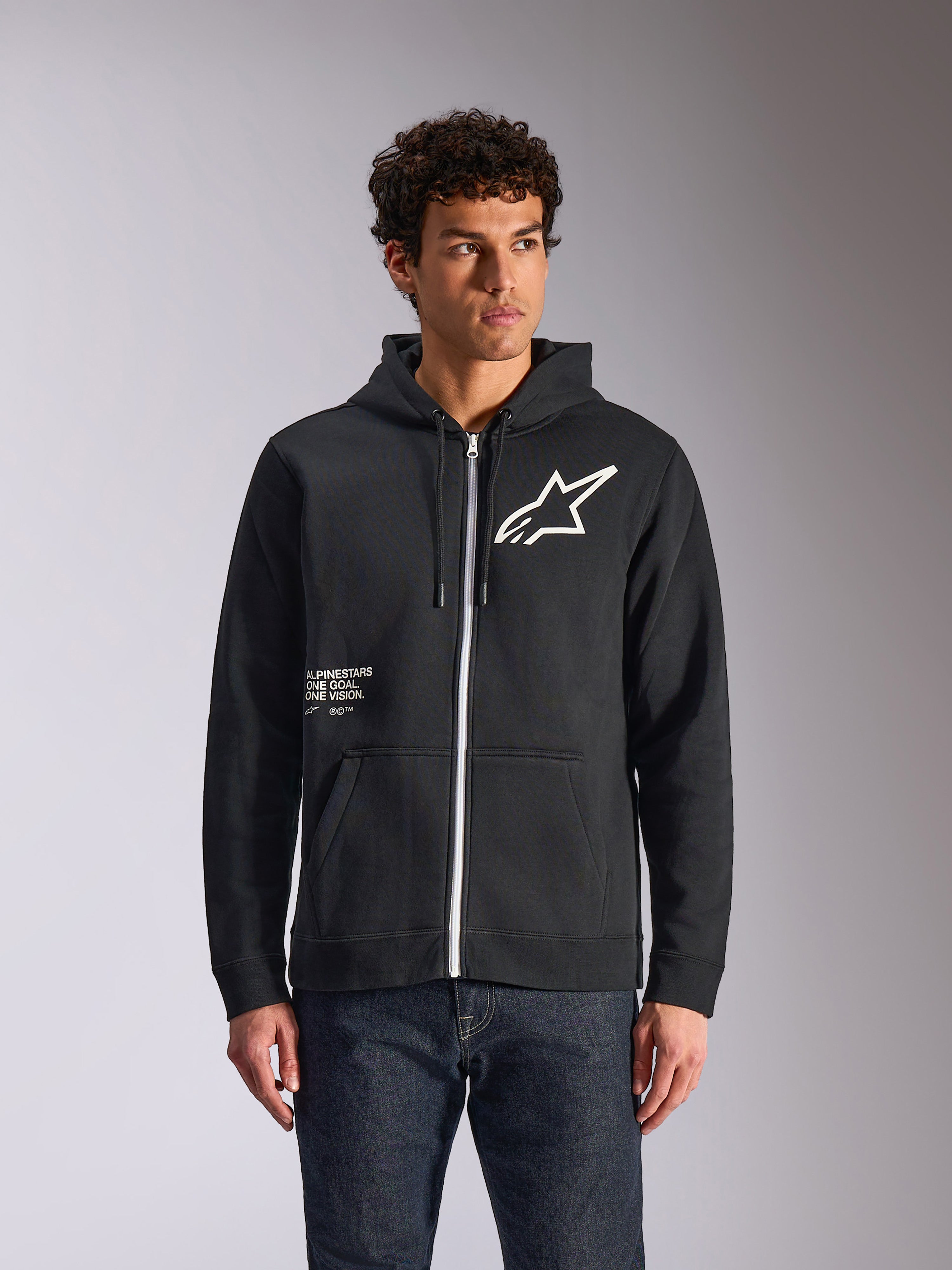 Alpinestars Transverse Hoodie, schwarzer Kapuzenfleece mit Reißverschluss vorne, weißes Astars-Logo und One Goal-Grafik auf der Vorderseite, getragen von einem Model mit dunkler Jeans, lässige, vom Motorsport inspirierte Streetwear