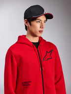 Alpinestars Transverse Hoodie, rotes Fleece mit Reißverschluss vorne, schwarzes Astars-Logo auf der Brust und „One Goal. One Vision.“-Schriftzug auf der unteren Vorderseite, lässige Motorsport-Streetwear, getragen von einem Model mit schwarzer Kappe