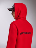 Alpinestars Transverse Hoodie, roter Kapuzenfleece, Seiten-Rückansicht mit schwarzem Alpinestars-Schriftzug auf dem Rücken, getragen mit hochgezogener Kapuze und einer schwarzen Kappe, lässige, vom Motorsport inspirierte Streetwear