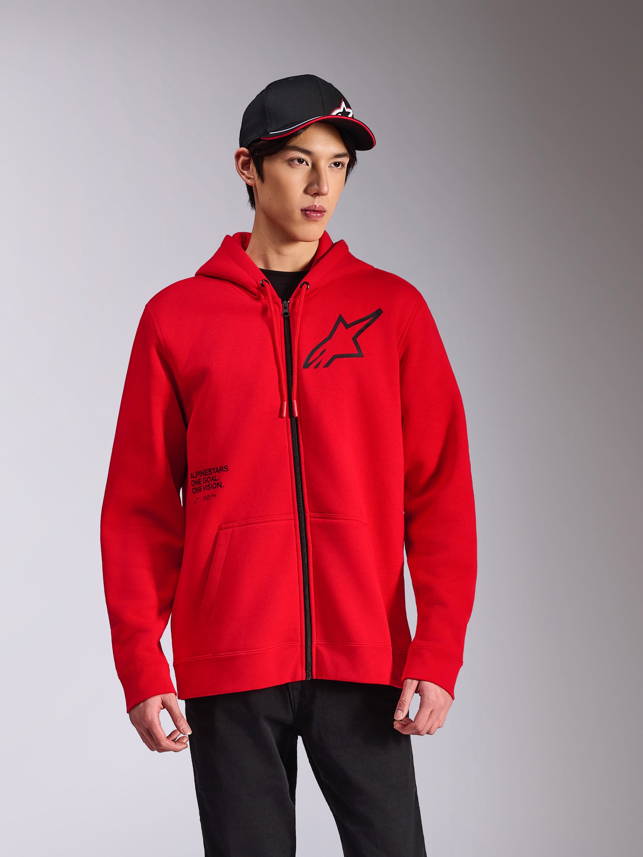 Alpinestars Transverse Hoodie, roter Fleece-Kapuzenpullover mit Frontreißverschluss, schwarzes Astars-Logo auf der Brust und „One Goal“-Aufdruck, lässiger Streetwear-Stil, getragen mit schwarzer Kappe und Hose