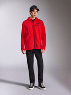 Alpinestars Transverse Hoodie, roter Fleece-Kapuzenpullover mit durchgehendem Reißverschluss, schwarzes Astars-Logo auf der Brust und One Goal-Druck an der Seite, getragen von einem männlichen Model mit schwarzer Hose und Kappe, lässige, vom Motorsport inspirierte Streetwear