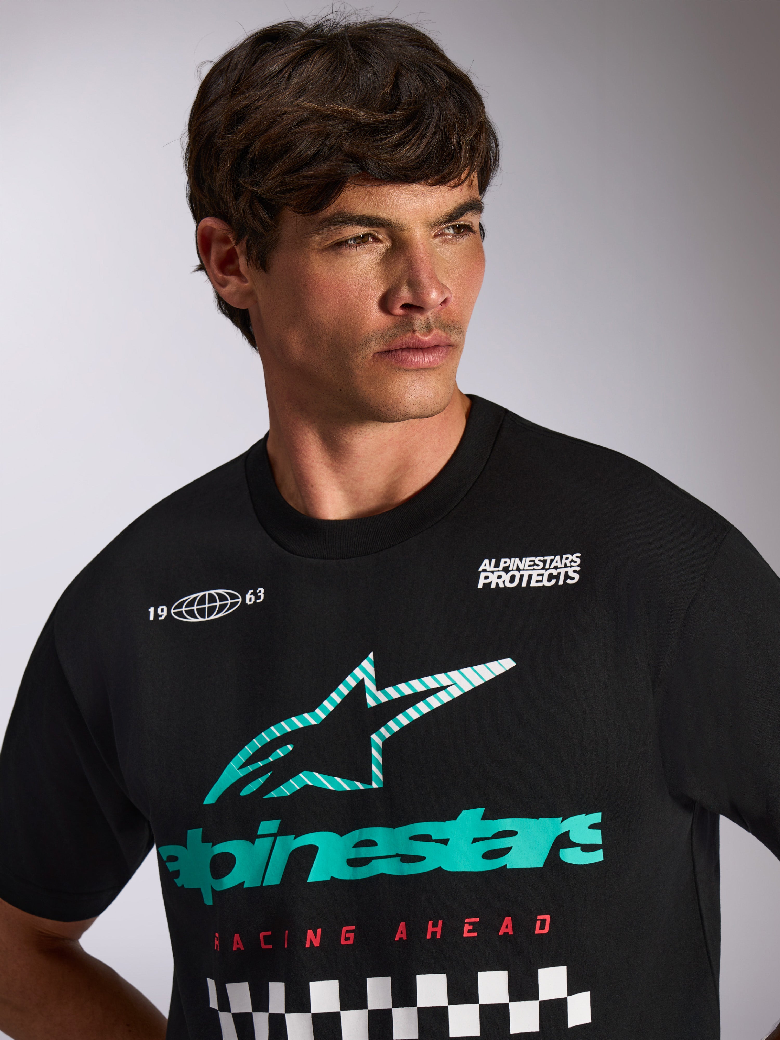 Alpinestars Aplomb Oversized CSF Tee, kurzärmeliges T-Shirt, Schwarz, türkis-weißes Astars-Kombi-Logo mit 'Racing Ahead'-Schriftzug und Zielflaggen-Muster, getragen von einem männlichen Model, lässige Motorsport-Streetwear