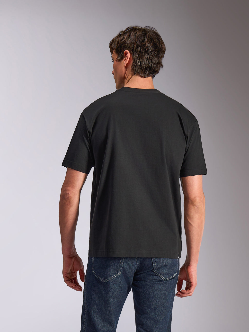 Alpinestars Aplomb Oversized CSF Tee, kurzärmeliges T-Shirt, Schwarz, Rückansicht, getragen von einem Model mit blauen Jeans, lockere Passform, lässige Sportbekleidung