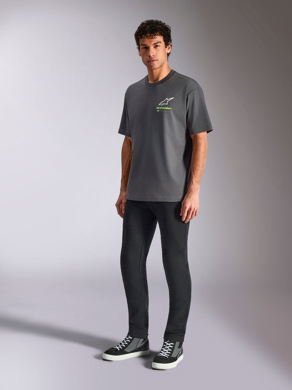 Alpinestars Overhang Oversized CSF Tee, Kurzarm-T-Shirt, Anthrazitgrau, mit weißem Astars-Logo und grünem Text auf der Brust, getragen von einem Model mit schwarzer Hose und Sneakern, lockere Oversized-Passform, legere Motorsport-Streetwear