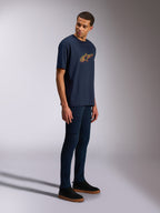 Alpinestars Alter Oversized CSF Tee, kurzärmeliges T-Shirt, Marineblau, gelbes und oranges Astars-Logo, getragen von einem Model mit dunklen Jeans und schwarzen Sneakern, lässige motorsport-inspirierte Streetwear