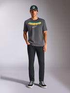 Alpinestars Ellipsoid CSF Tee, Kurzarm-T-Shirt, Anthrazitgrau, mit einem geschwungenen gelb-orangen Farbverlauf-Schriftzug und weißem Astars-Logo, getragen von einem Model mit schwarzer Hose und Baseballkappe, lässiger Sportbekleidungsstil