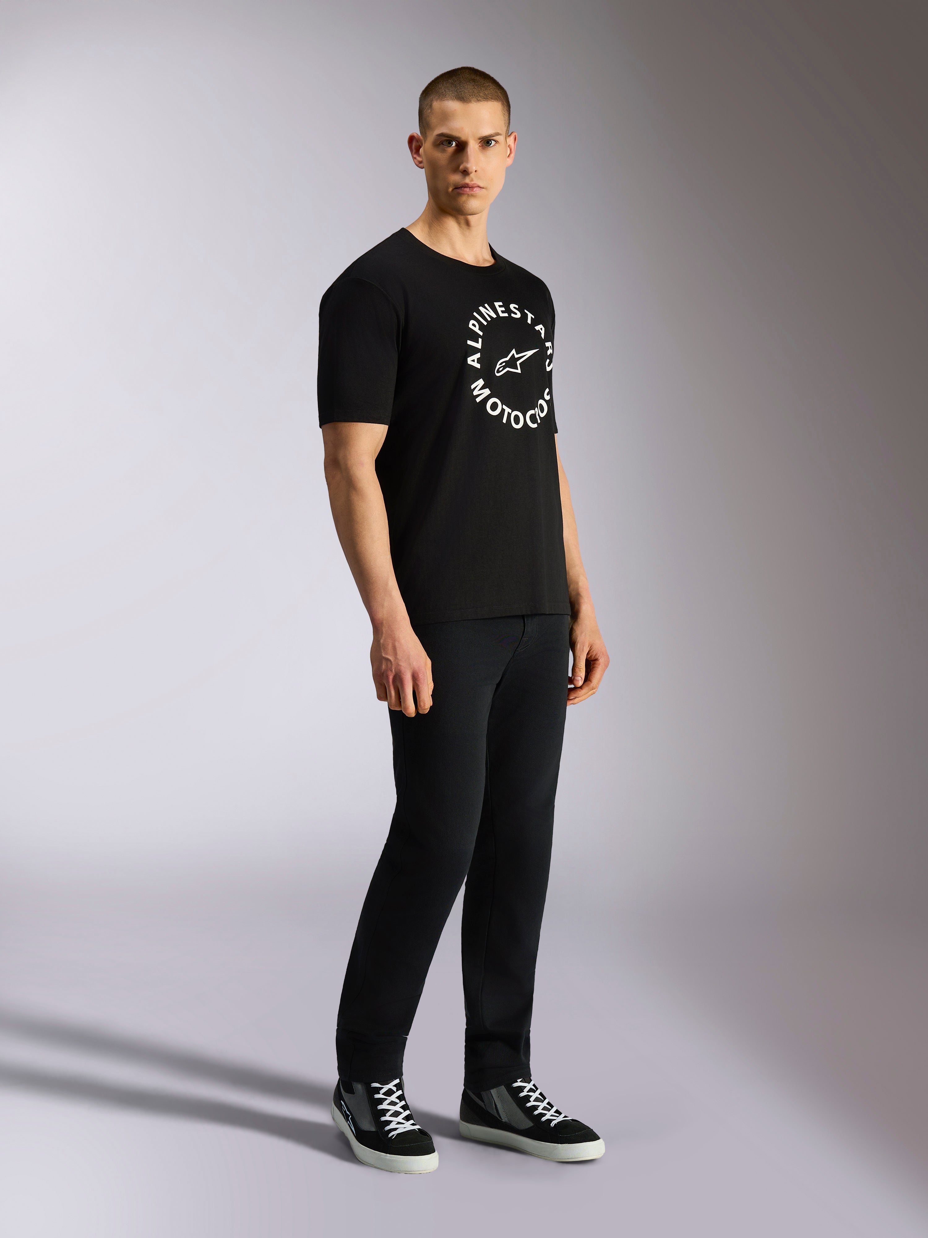 Alpinestars MX CSF Tee, Kurzarm-T-Shirt, Schwarz, weiße kreisförmige Alpinestars Motocross-Grafik auf der Brust, von einem Model mit schwarzer Hose und Sneakern getragen, lässige Motorsport-Streetwear