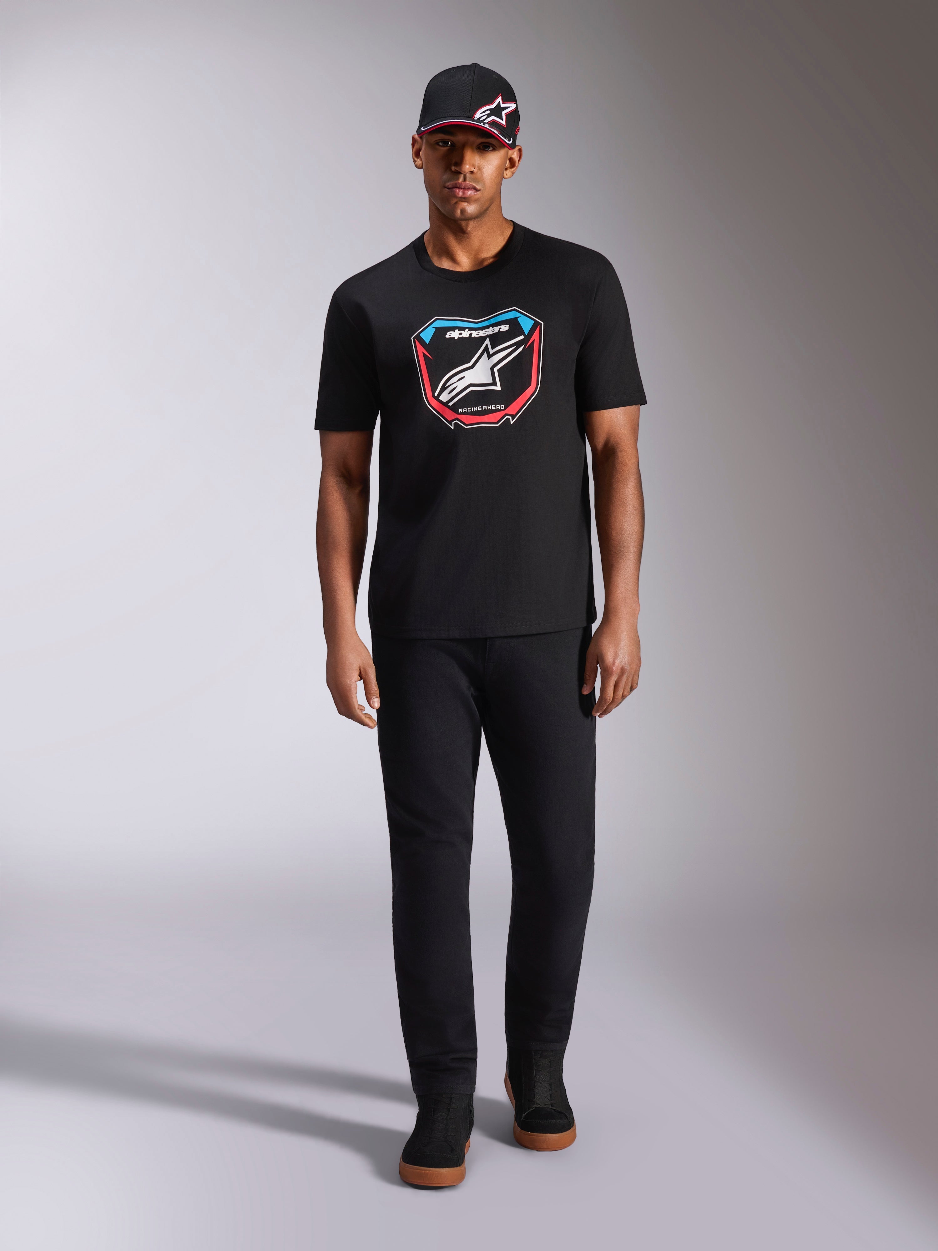 Alpinestars Plate CSF T-Shirt, Kurzarm, Schwarz, MX-Plattengrafik auf der Vorderseite mit weißem Astars-Logo und blauen sowie roten Akzenten, getragen von einem Model mit passender schwarzer Kappe und Hose, lässige Motorsport-Sportbekleidung