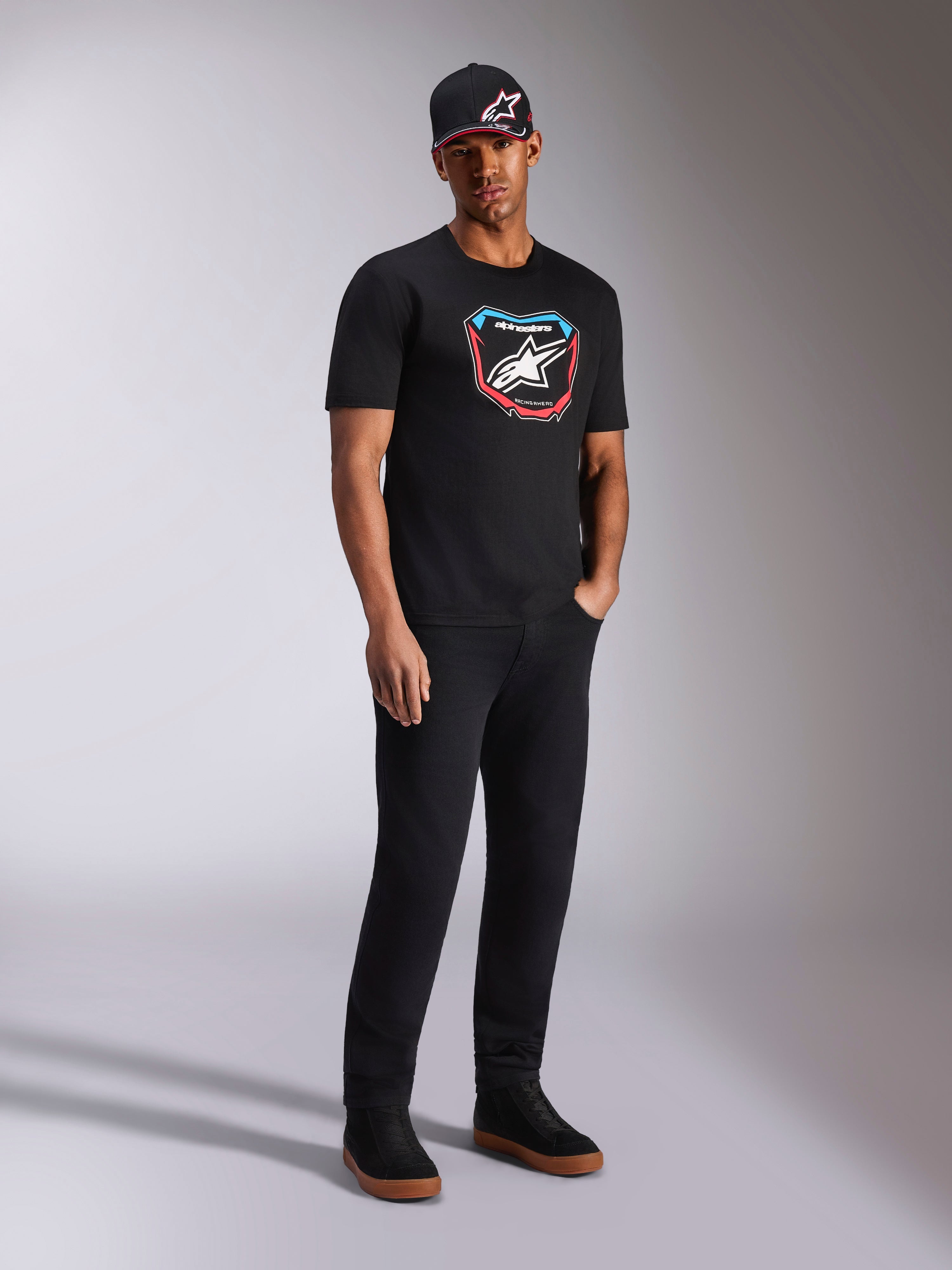 Alpinestars Plate CSF Tee, Kurzarm-T-Shirt, Schwarz, bunte MX-Plattengrafik auf der Vorderseite mit Stern-Logo, getragen von einem Model mit schwarzer Logo-Kappe und Hose, lässige, vom Motorsport inspirierte Streetwear