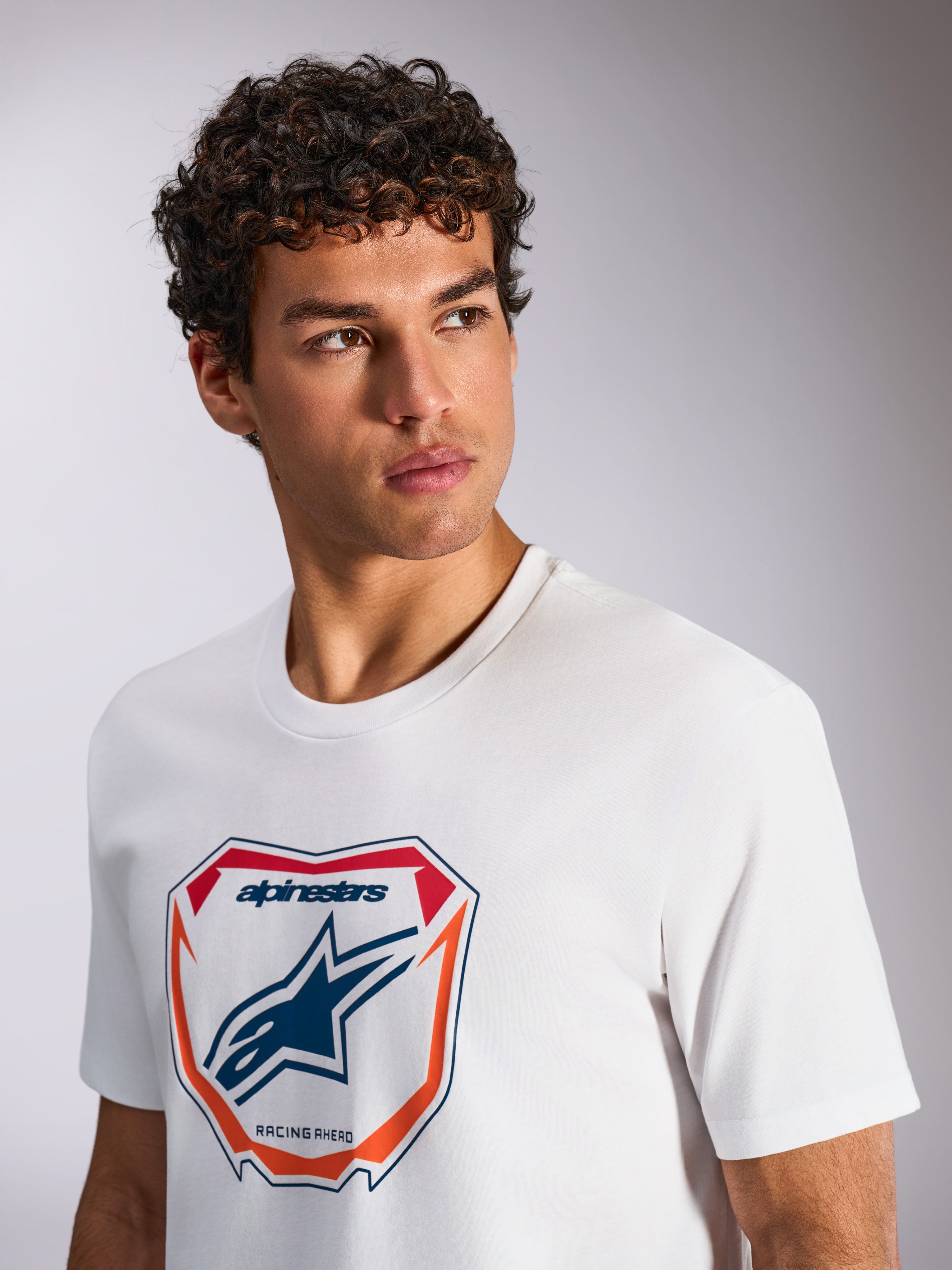 Alpinestars Plate CSF Tee, Kurzarm-T-Shirt, Weiß, mit einer Astars MX Plattengrafik auf der Vorderseite mit roten und orangen Akzenten, „Racing Ahead“-Schriftzug, von einem Model getragen, lässige Motorsport-Streetwear
