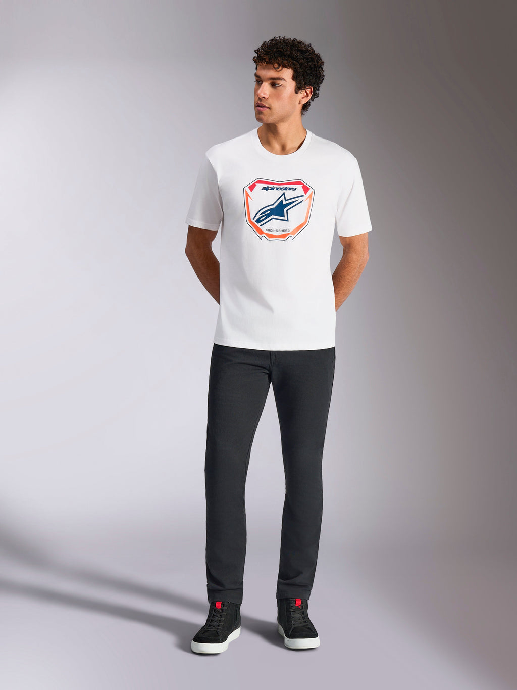 Alpinestars Plate CSF Tee, Kurzarm-T-Shirt, Weiß, mit einer Astars MX-Plattengrafik auf der Vorderseite mit orangefarbenen und blauen Akzenten, getragen von einem Model mit schwarzer Hose und Sneakern, legere Motorsport-Streetwear