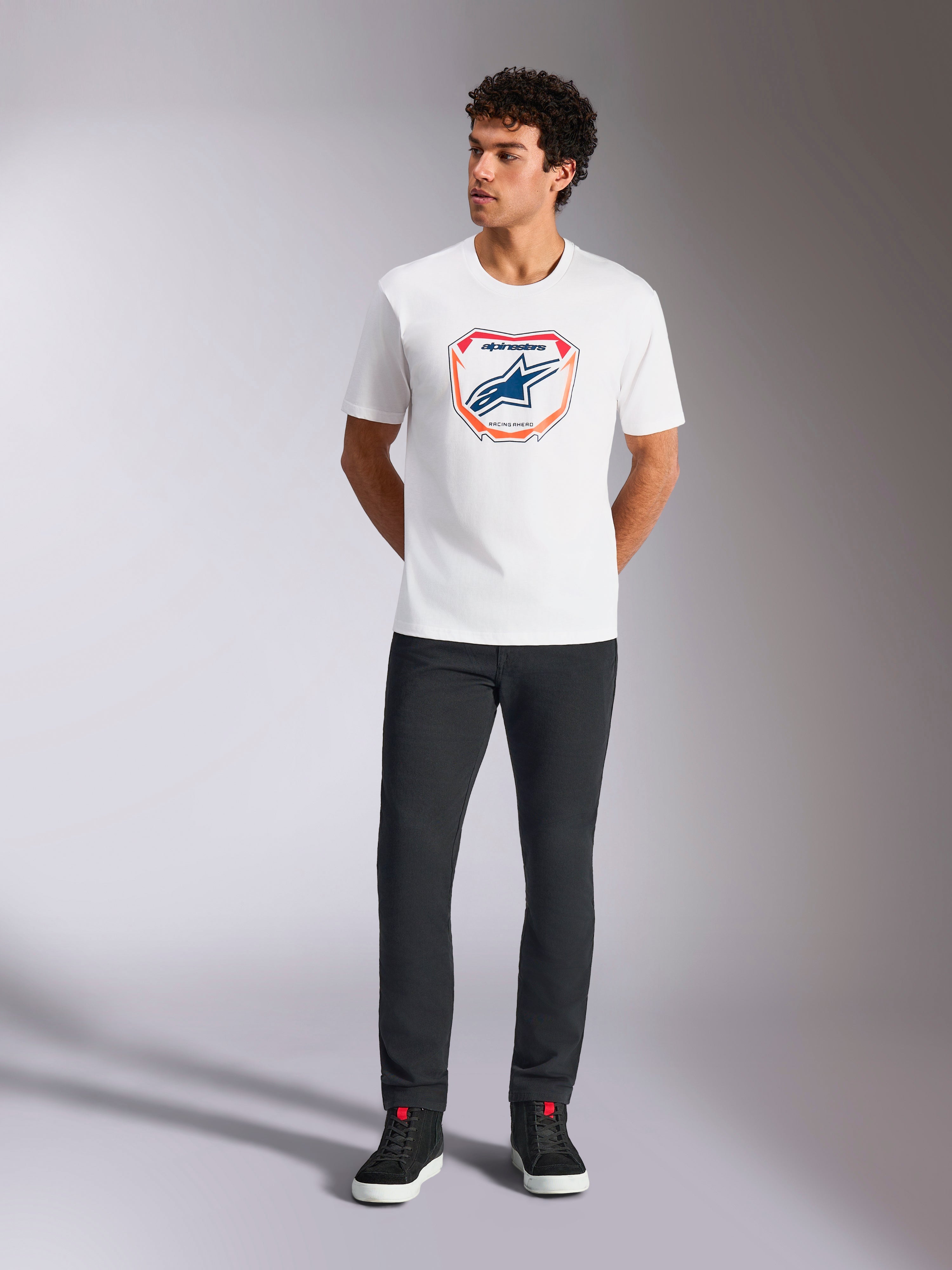 Alpinestars Plate CSF Tee, Kurzarm-T-Shirt, Weiß, mit einer Astars MX-Plattengrafik auf der Vorderseite mit orangefarbenen und blauen Akzenten, getragen von einem Model mit schwarzer Hose und Sneakern, legere Motorsport-Streetwear