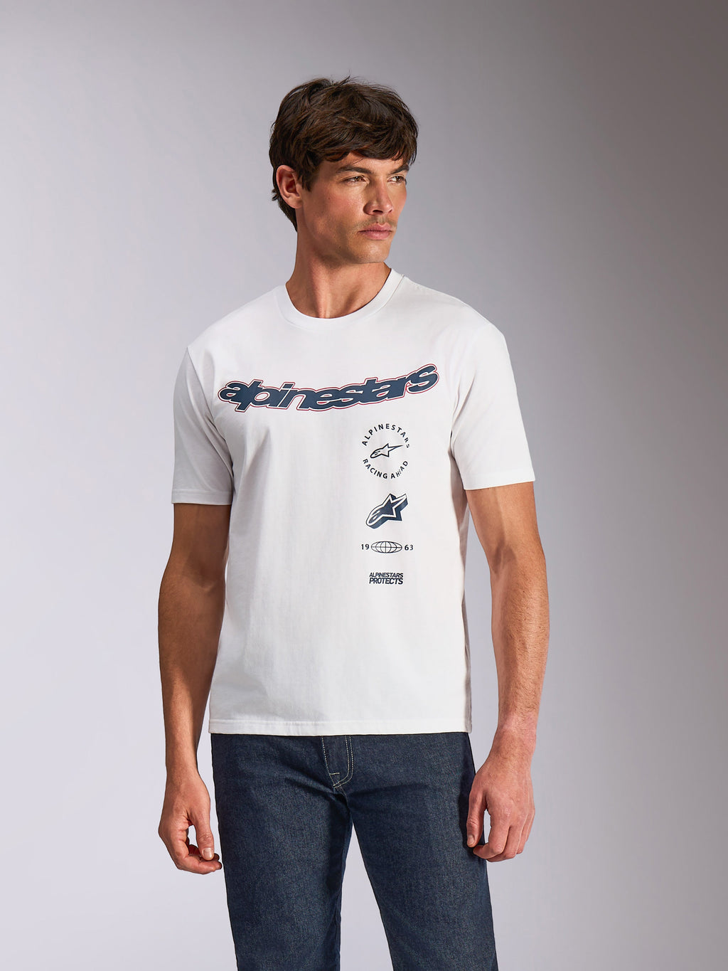 Alpinestars Abus CSF Tee, kurzärmeliges T-Shirt, Weiß, geschwungenes Alpinestars-Logo mit mehreren gestapelten Grafiken auf der Brust, getragen von einem Model mit dunklen Denim-Jeans, lässige Motorsport-Streetwear