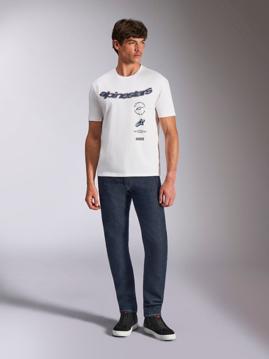 Alpinestars Abus CSF Tee, kurzärmeliges T-Shirt, Weiß, mit geschwungenem Alpinestars-Logo und mehreren übereinander angeordneten Grafiken auf der Brust, getragen von einem männlichen Model mit dunkelblauer Jeans und schwarzen Sneakern, lässige Motorsport-Streetwear