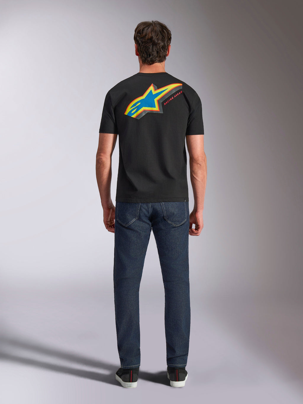 Alpinestars Angler CSF Tee, Kurzarm-T-Shirt, Schwarz, Rückansicht eines Models im Shirt mit großem, leuchtendem, mehrfarbigem Astars-Sternlogo und „RACING AHEAD“-Text, kombiniert mit dunkelblauen Jeans, lässige Motorsport-Streetwear