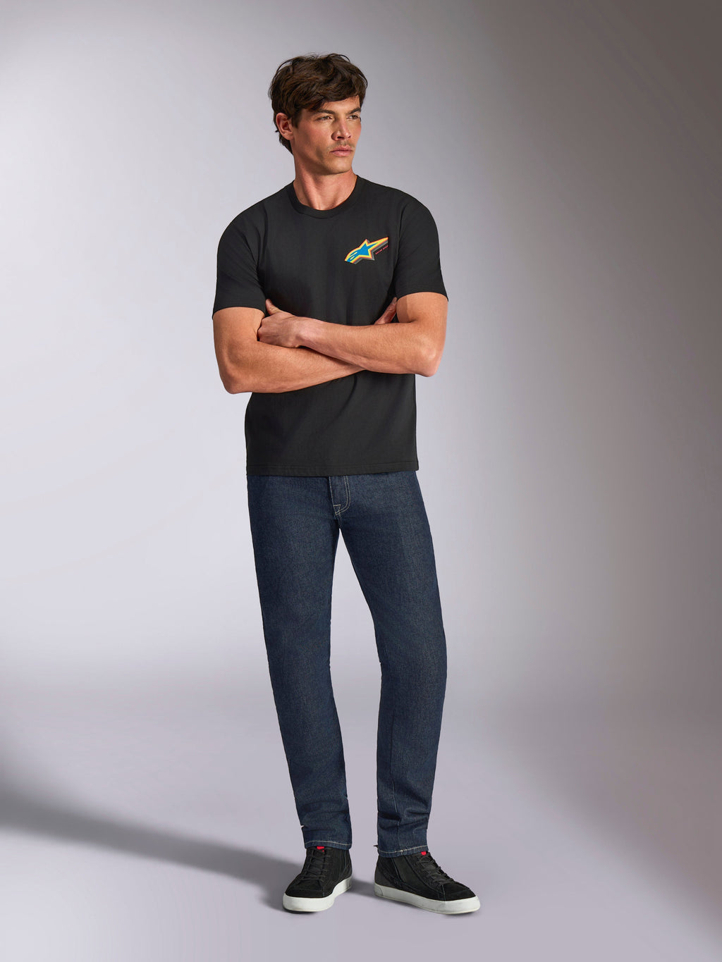 Alpinestars Angler CSF Tee, kurzärmeliges T-Shirt, Schwarz, lebendiges mehrfarbiges Astars-Logo auf der Brust, Modell mit verschränkten Armen, dunkle Denim-Jeans und schwarze Sneaker, lässige Motorsport-Streetwear