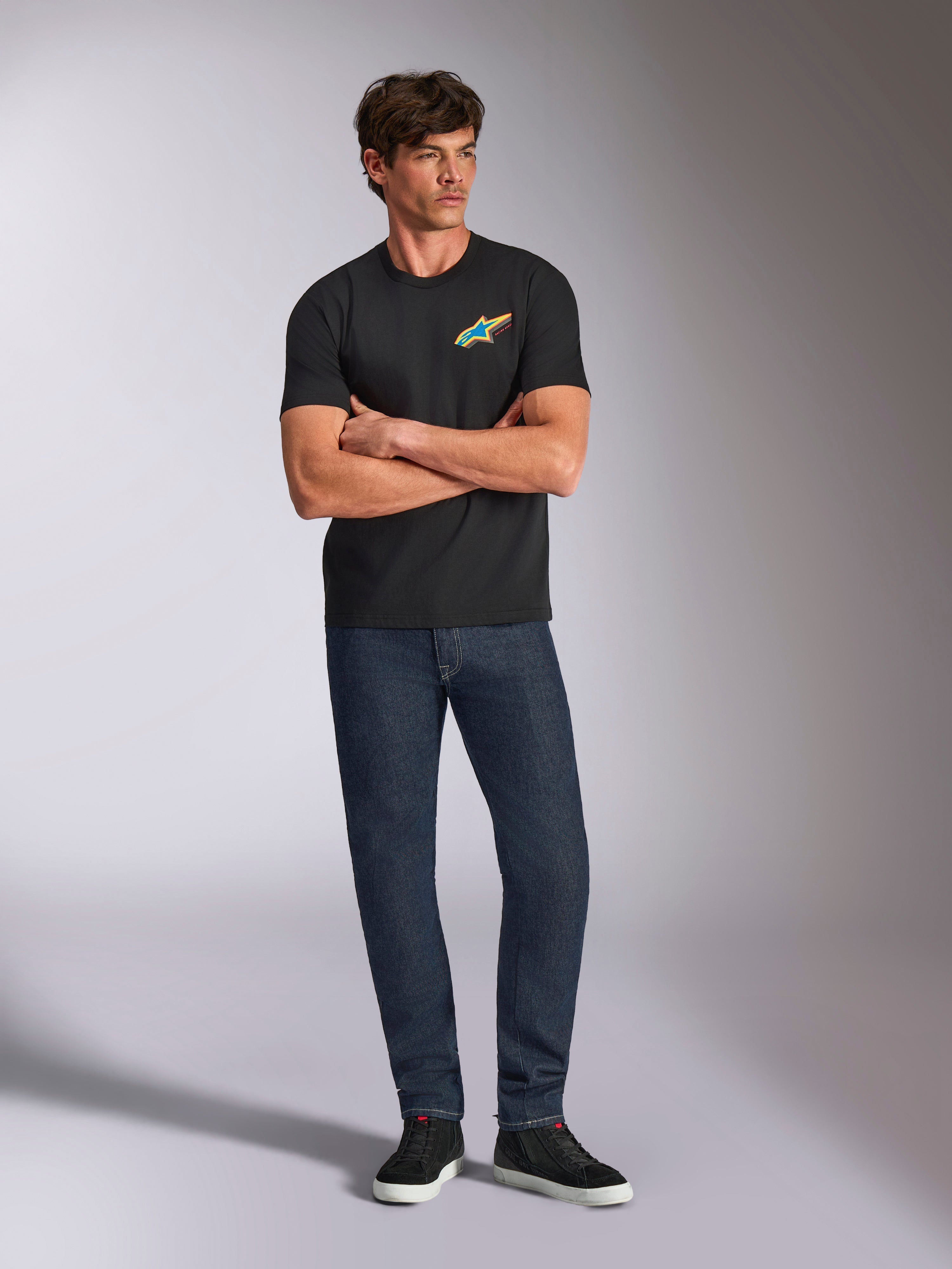 Alpinestars Angler CSF Tee, kurzärmeliges T-Shirt, Schwarz, lebendiges mehrfarbiges Astars-Logo auf der Brust, Modell mit verschränkten Armen, dunkle Denim-Jeans und schwarze Sneaker, lässige Motorsport-Streetwear
