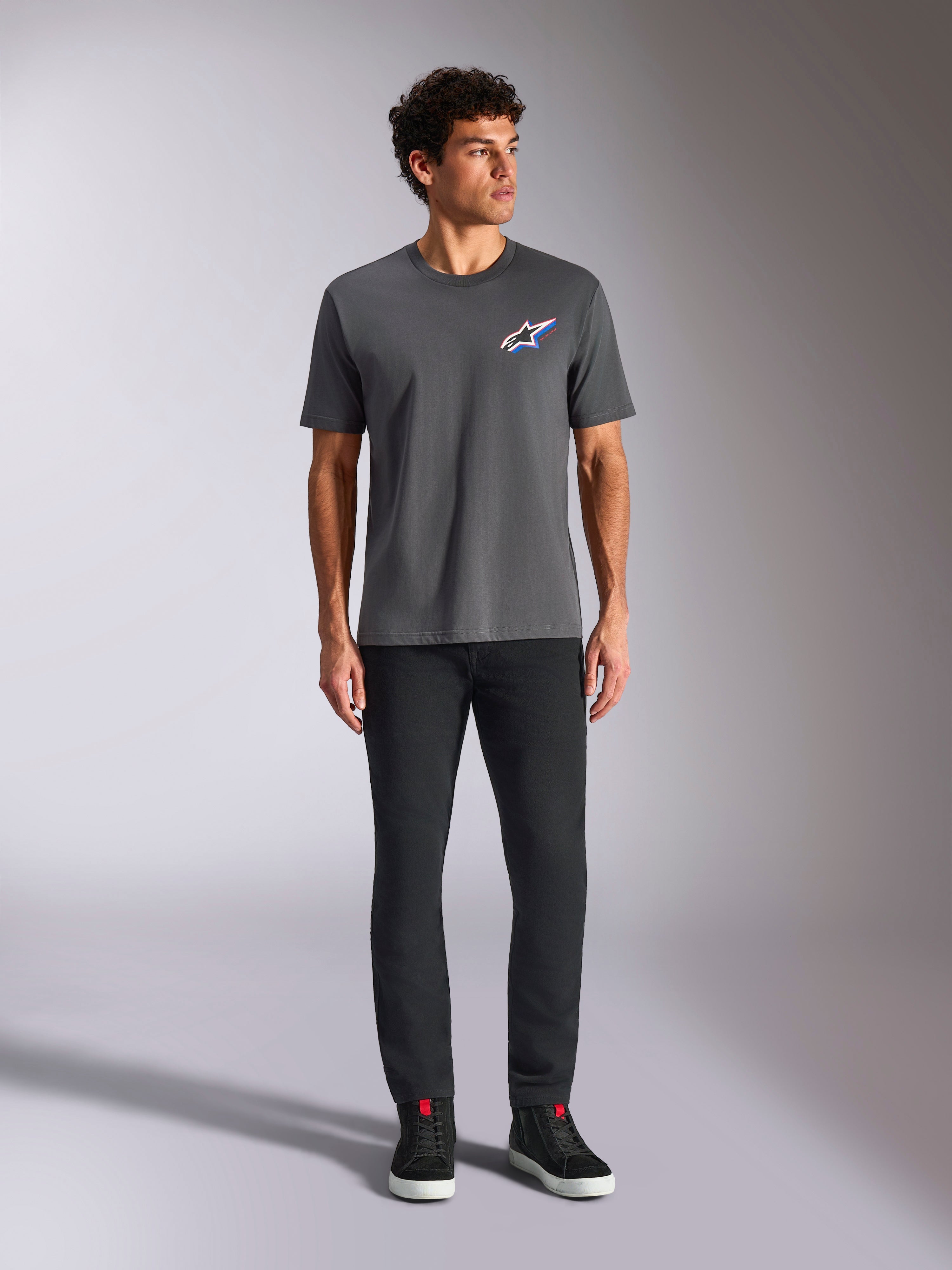 Alpinestars Angler CSF Tee, Kurzarm-T-Shirt, Anthrazit, mit einem lebendigen mehrfarbigen Astars-Logo auf der Brust, getragen von einem Model mit schwarzer Hose und Sneakern, Freizeit-Motorsport-Streetwear