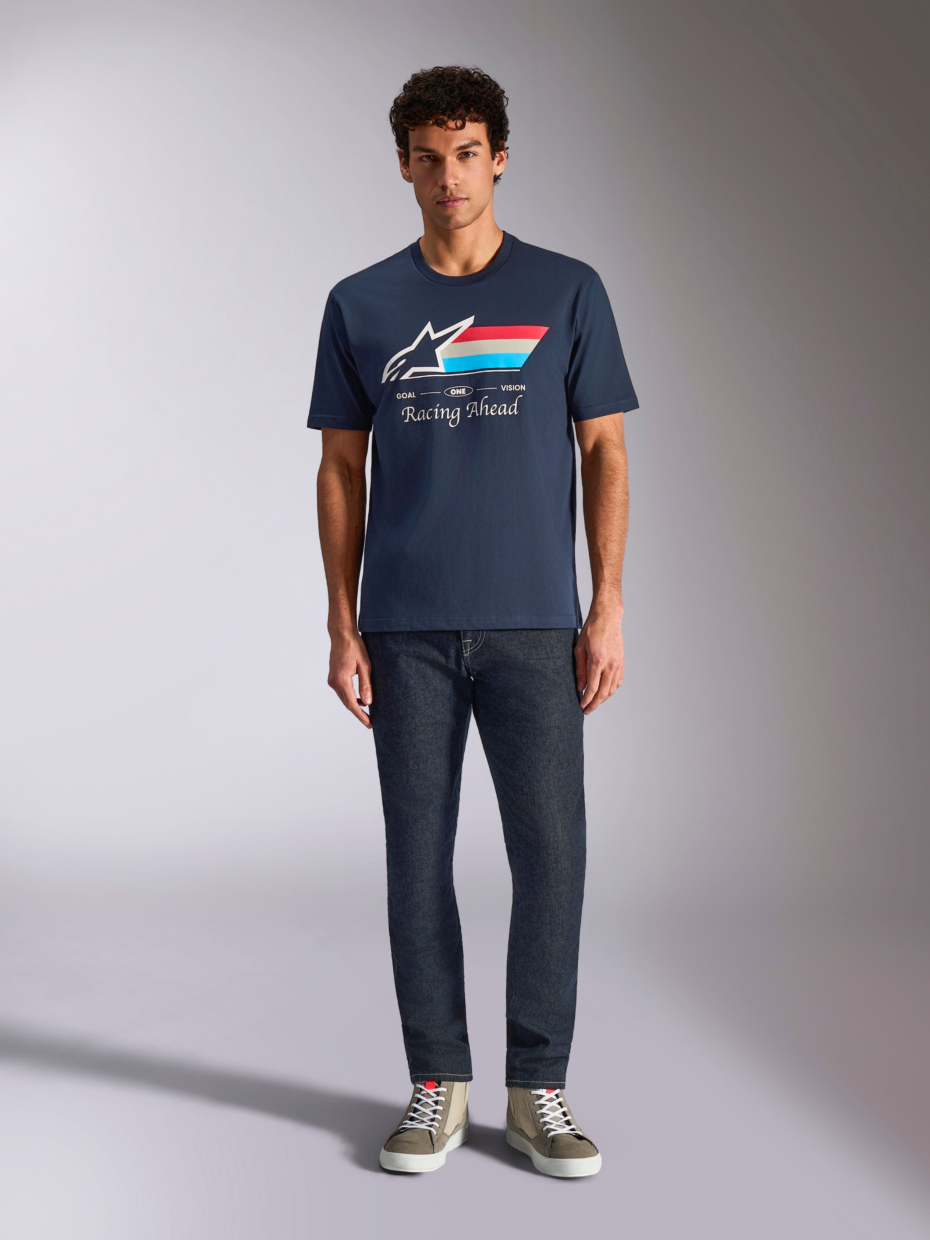Alpinestars Trifecta CSF Tee, Kurzarm-T-Shirt, Marineblau, weißes Astars-Logo mit rot-weiß-hellblauen Streifen und „Racing Ahead“-Schriftzug, getragen mit dunkler Jeans und olivgrünen Sneakern, lässige Motorsport-Streetwear