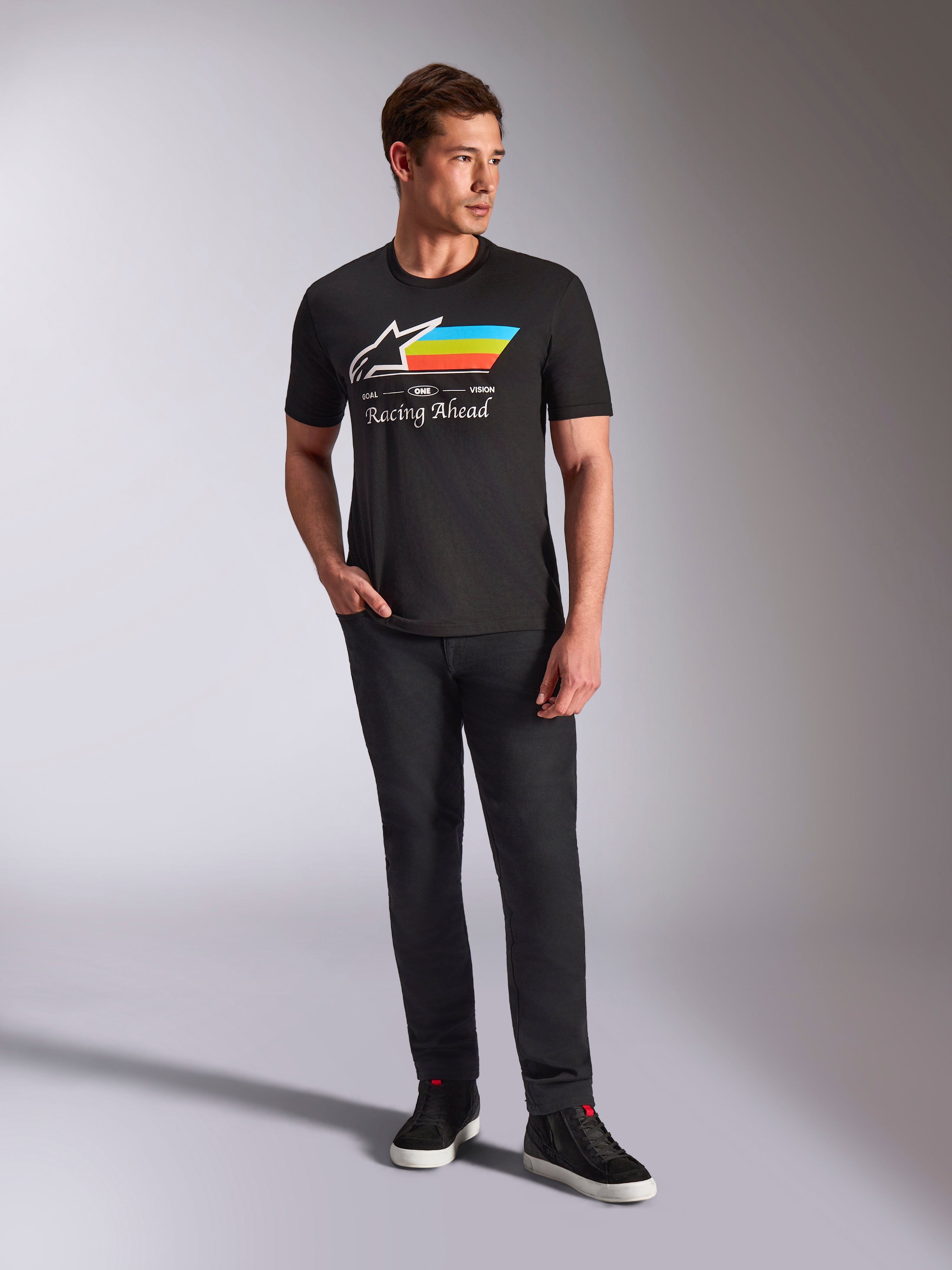 Alpinestars Trifecta CSF Tee, Short Sleeve T-Shirt, Black, featuring a white Astars logo with cyan, yellow, and orange stripes and 'Racing Ahead' text, worn by a model with black pants and sneakers for casual motorsport streetwear