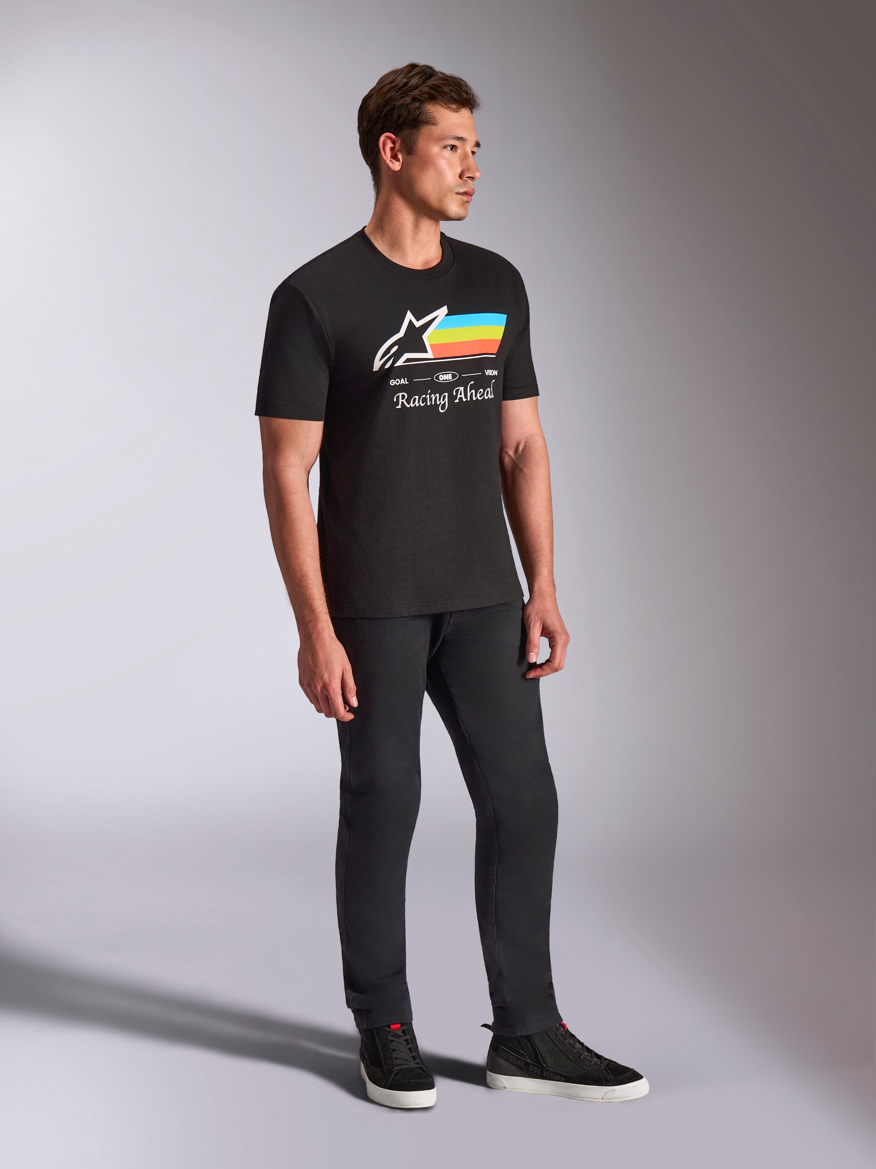 Alpinestars Trifecta CSF Tee, Short Sleeve T-Shirt, Black, white Astars logo with blue, yellow, and orange stripes and 'Racing Ahead' graphic, worn by a model with black pants and sneakers, casual motorsport-inspired lifestyle apparel