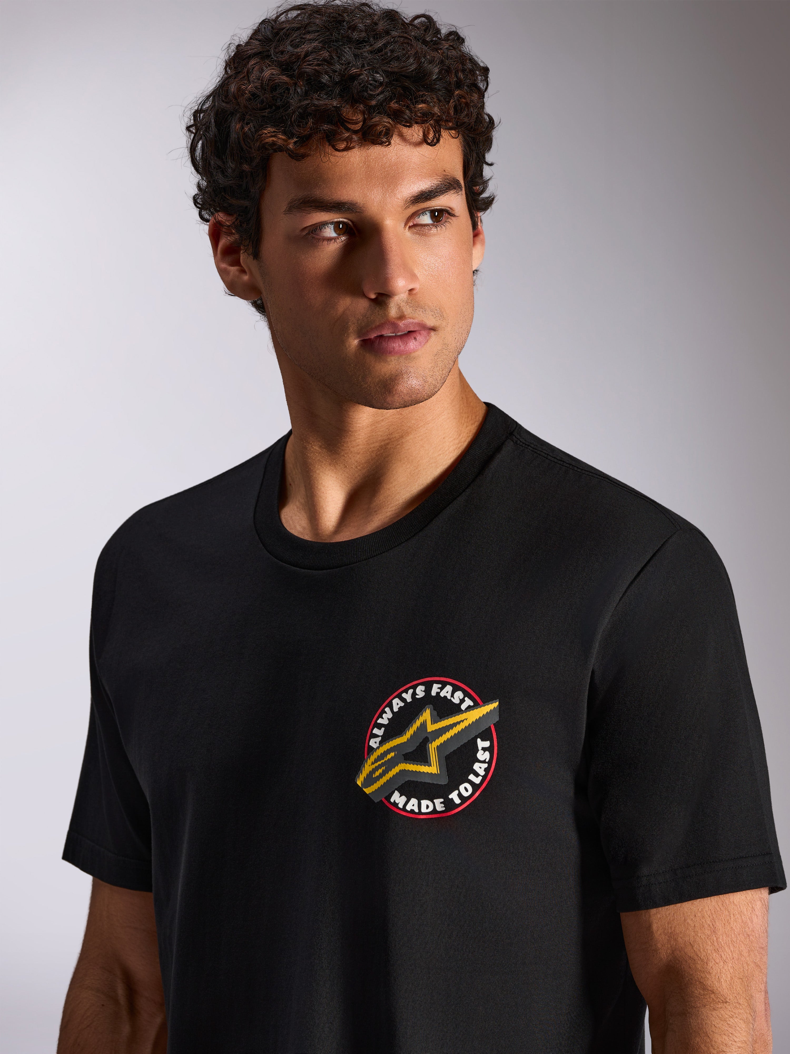 Alpinestars Aorbit CSF Tee, kurzärmeliges T-Shirt, Schwarz, kreisförmige „Always Fast Made to Last“-Brustgrafik mit gelbem Astars-Logo, von einem Model getragen, Casual Motorsport Streetwear