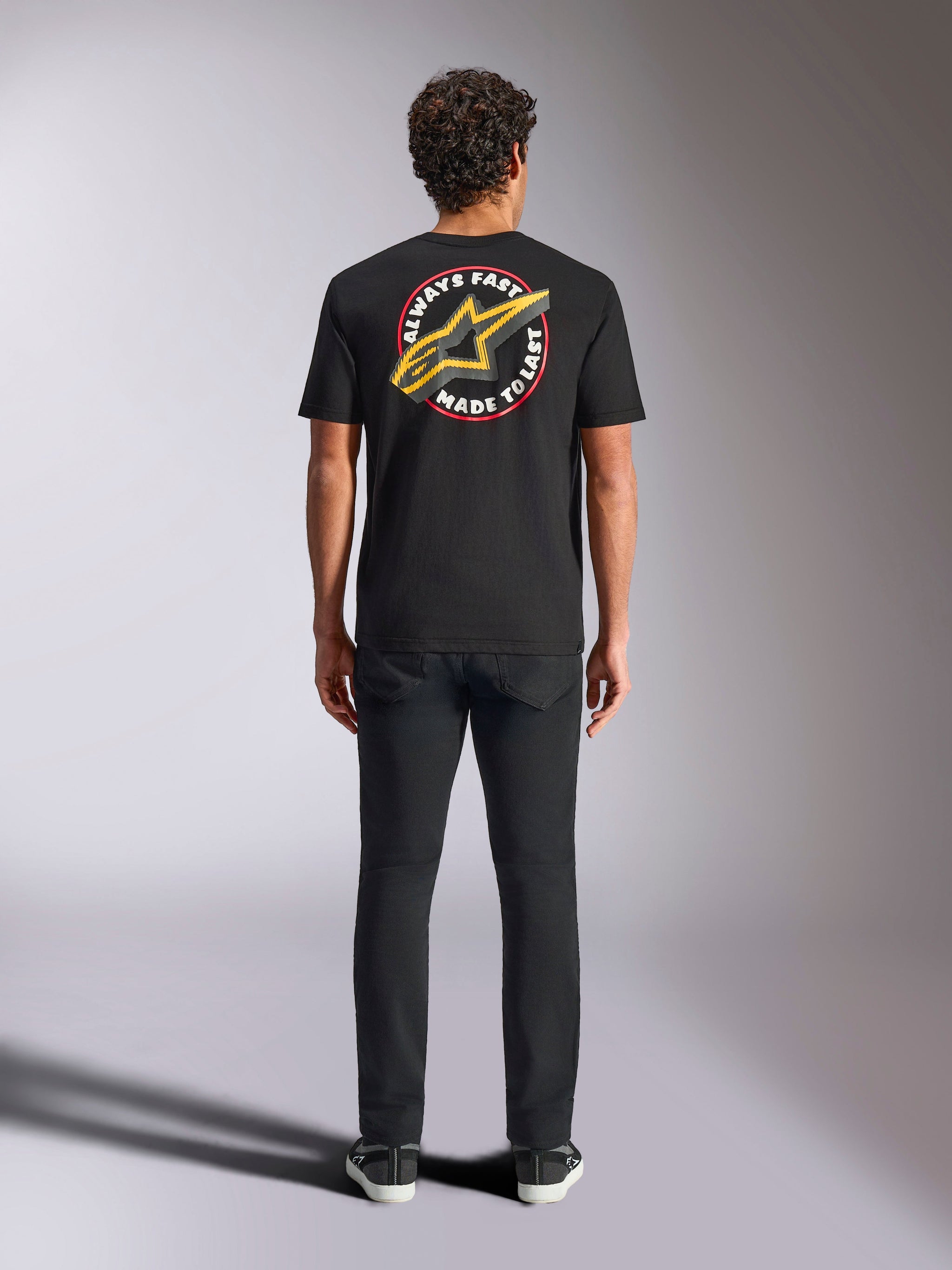 Alpinestars Aorbit CSF Tee, Short Sleeve T-Shirt, Black, worn by model from the back with black pants, featuring large 'Always Fast Made to Last' graphic with yellow Astars logo, casual motorsport streetwear
