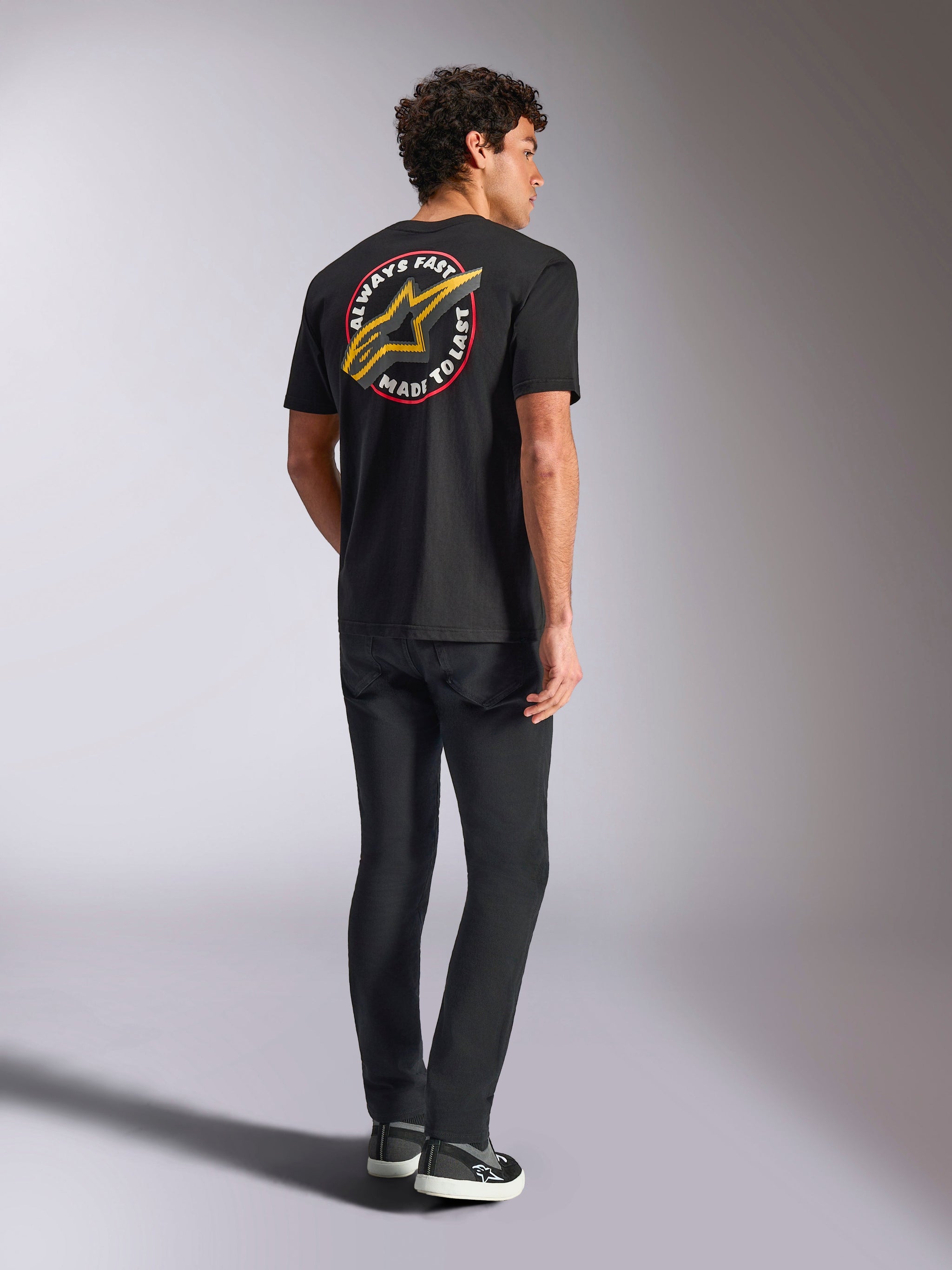 Alpinestars Aorbit CSF Tee, Short Sleeve T-Shirt, Black, back view featuring a large yellow Astars logo with 'Always Fast Made to Last' circular graphic, worn with black jeans and casual sneakers, motorsport-inspired streetwear style