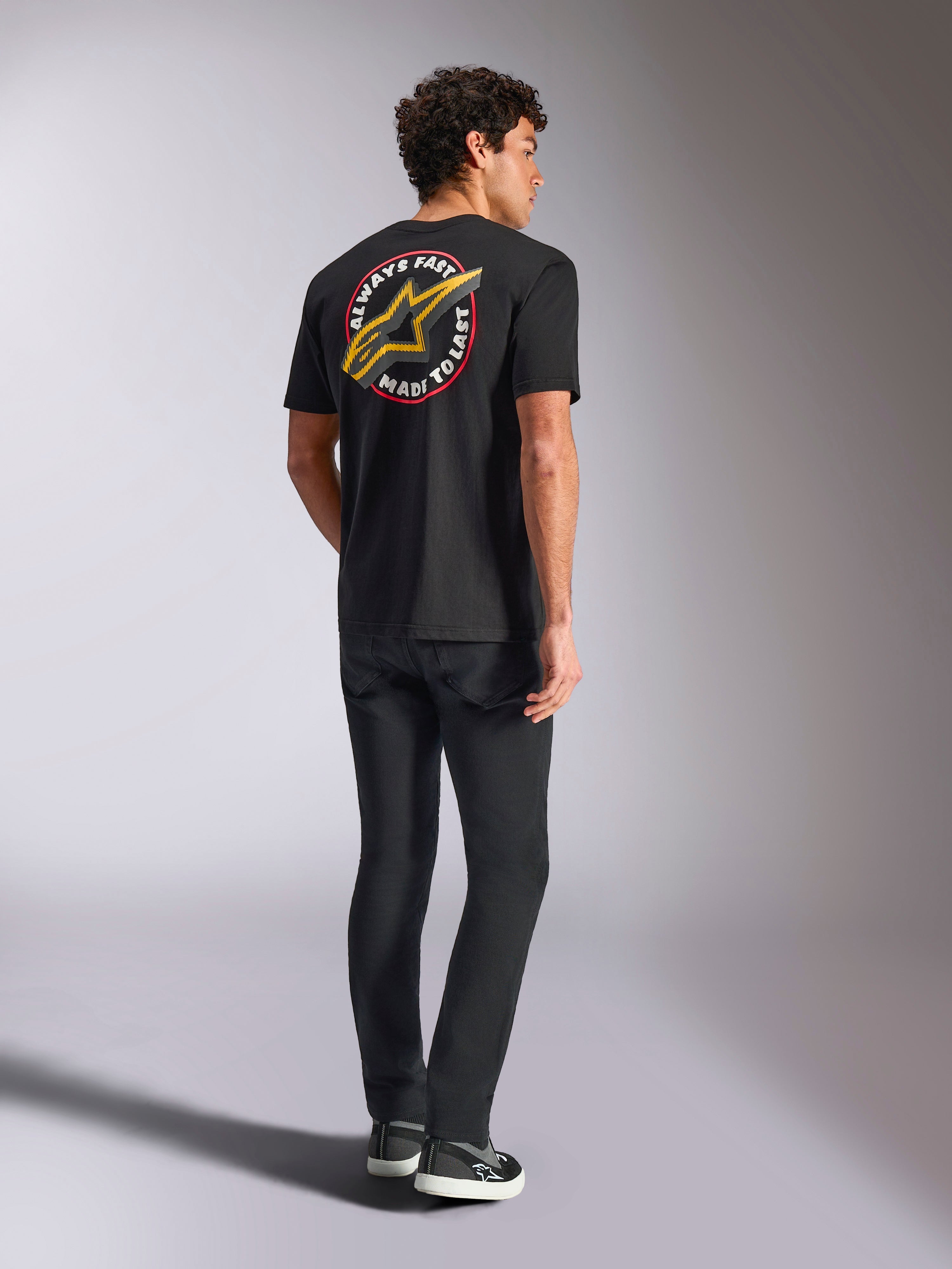 Alpinestars Aorbit CSF Tee, Kurzarm-T-Shirt, Schwarz, Rückansicht mit einem großen gelben Astars-Logo und kreisförmiger „Always Fast Made to Last“-Grafik, getragen mit schwarzen Jeans und lässigen Sneakern, Motorsport-inspirierter Streetwear-Stil