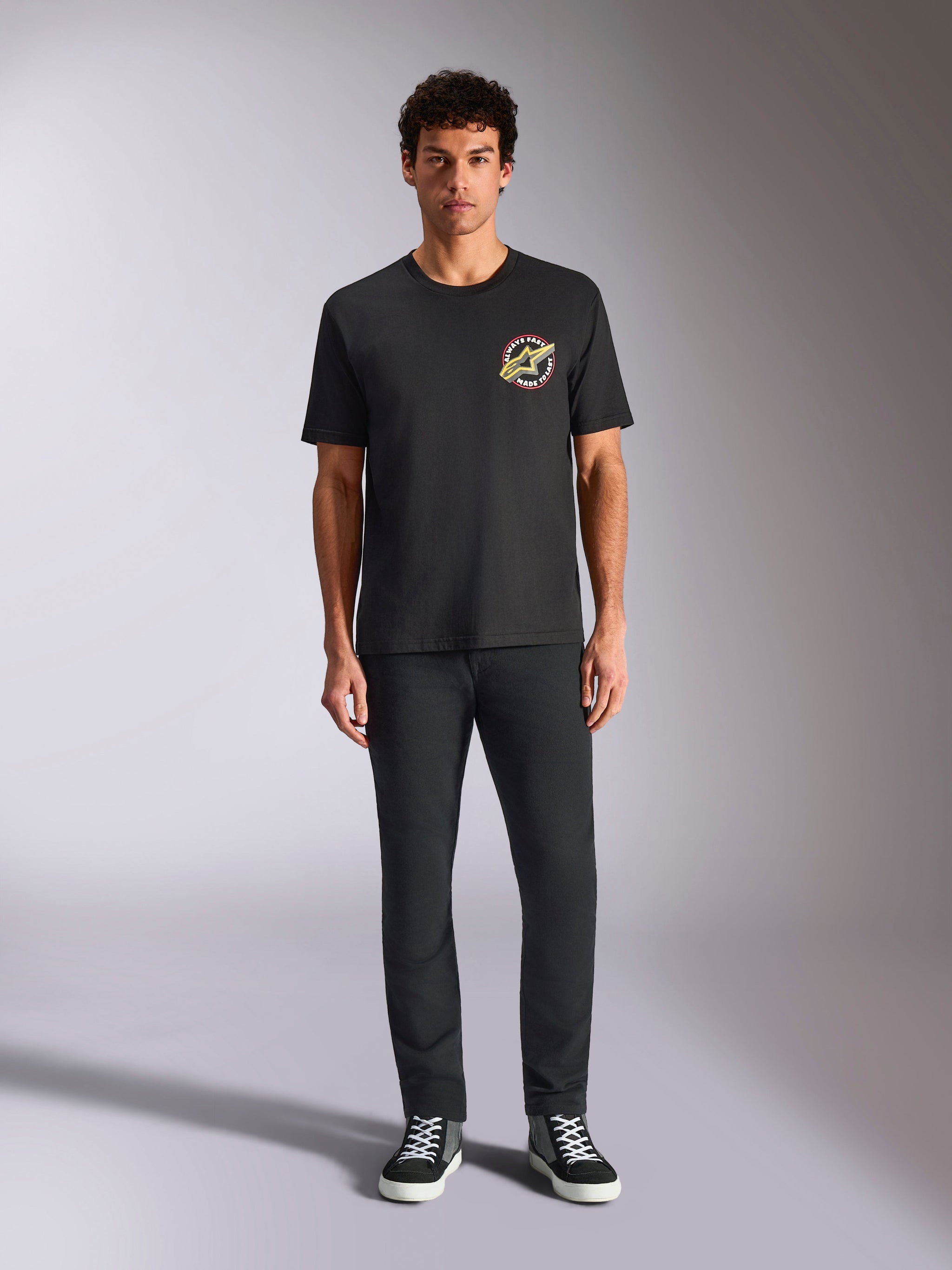 Alpinestars Aorbit CSF Tee, Short Sleeve T-Shirt, Black, circular logo with 'Always Fast Made to Last' text on the chest, worn by a model with black pants and sneakers, casual motorsport-inspired streetwear