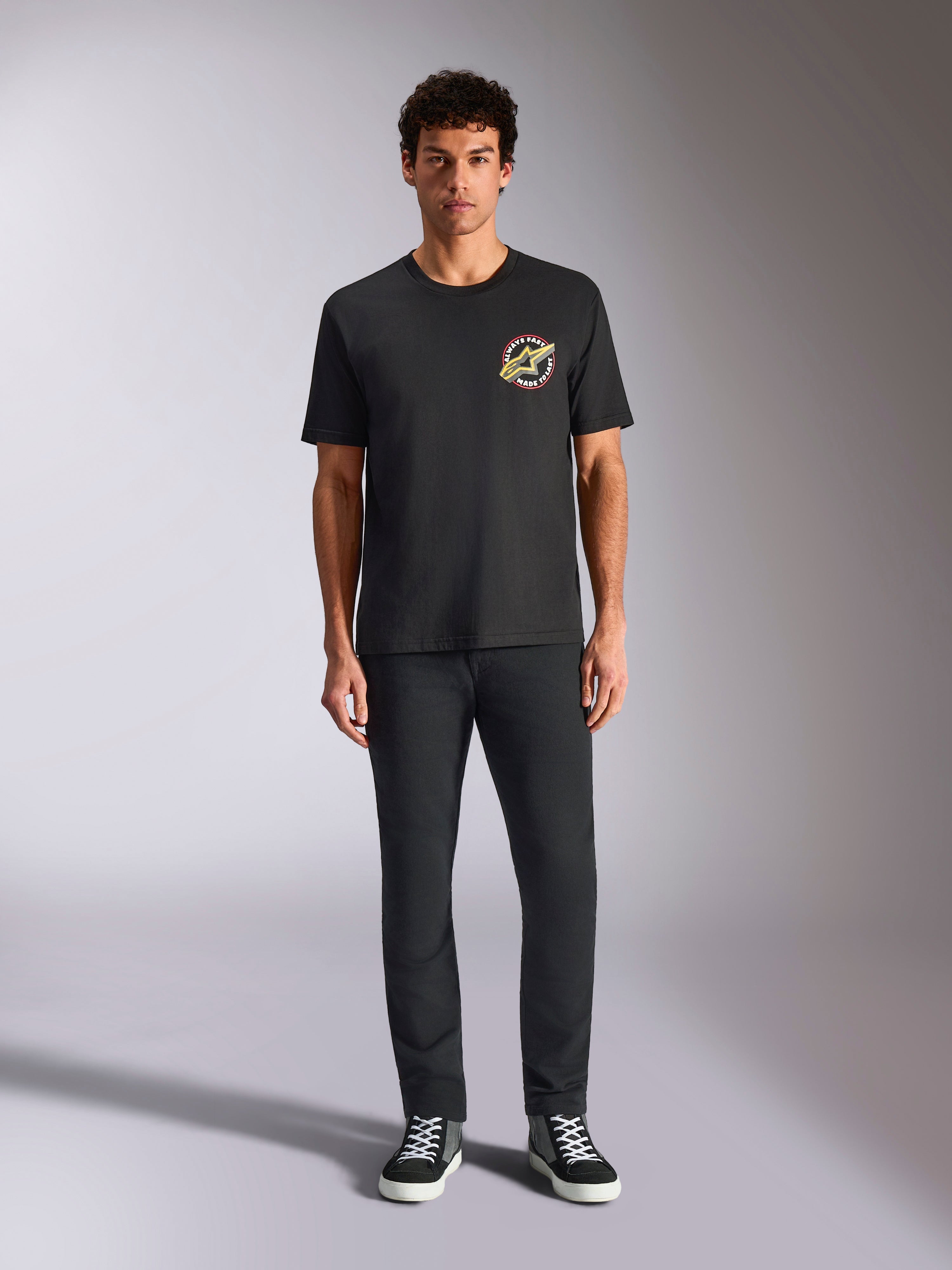 Alpinestars Aorbit CSF Tee, Kurzarm-T-Shirt, Schwarz, kreisförmiges Logo mit „Always Fast Made to Last“-Schriftzug auf der Brust, getragen von einem Model mit schwarzer Hose und Sneakern, lässige, vom Motorsport inspirierte Streetwear