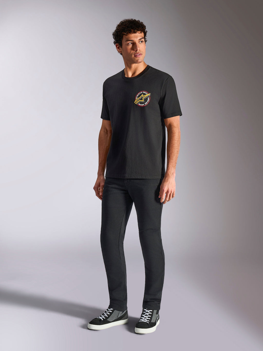 Alpinestars Aorbit CSF Tee, kurzärmeliges T-Shirt, Schwarz, mit kreisförmiger „Always Fast Made to Last“-Grafik auf der Brust, getragen von einem Model mit dunkler schmaler Hose und Sneakern, lässige Motorsport-Streetwear