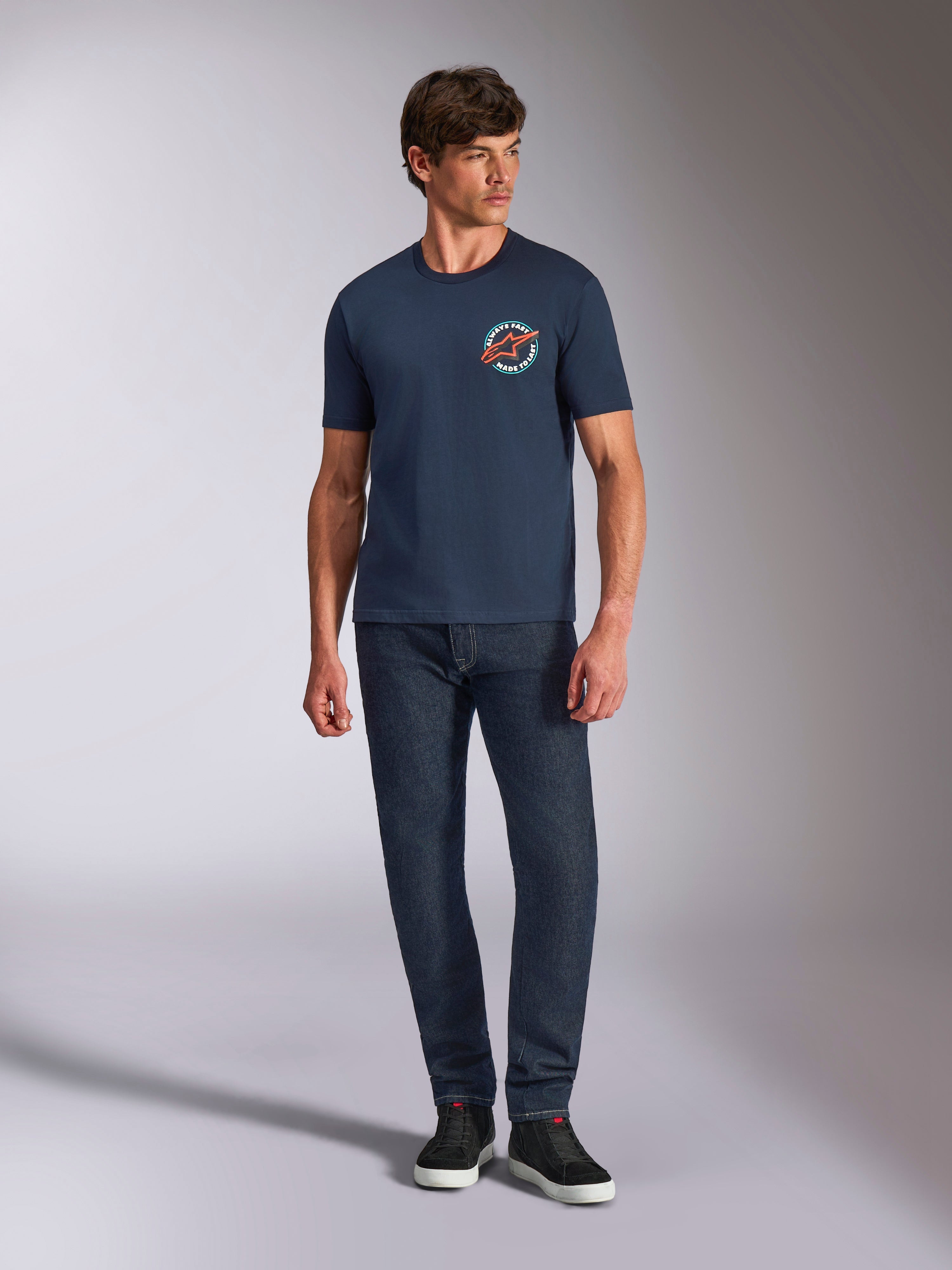 Alpinestars Aorbit CSF Tee, kurzärmeliges T-Shirt, Navy, kreisförmige Brustgrafik mit „Always Fast Made to Last“-Text, getragen mit dunkler Jeans und Sneakern, lässige Motorsport-Lifestyle-Bekleidung
