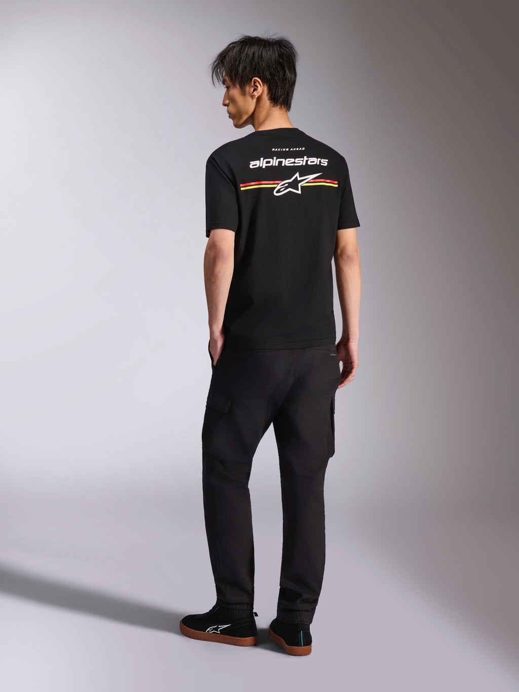 Alpinestars Lanes CSF Tee, schwarzes Kurzarm-T-Shirt, Rückansicht mit weißem Alpinestars-Logo sowie roten, orangen und gelben Streifengrafiken, lässige Motorsport-Streetwear kombiniert mit einer schwarzen Cargo-Hose