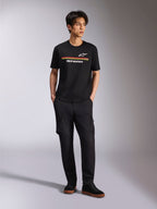 Model trägt Alpinestars Lanes CSF Tee, Kurzarm-T-Shirt, Schwarz, mit roten und gelben Streifen sowie Astars- und Alpinestars-Logos, kombiniert mit schwarzer Cargo-Hose und Sneakern, lässige Motorsport-Streetwear